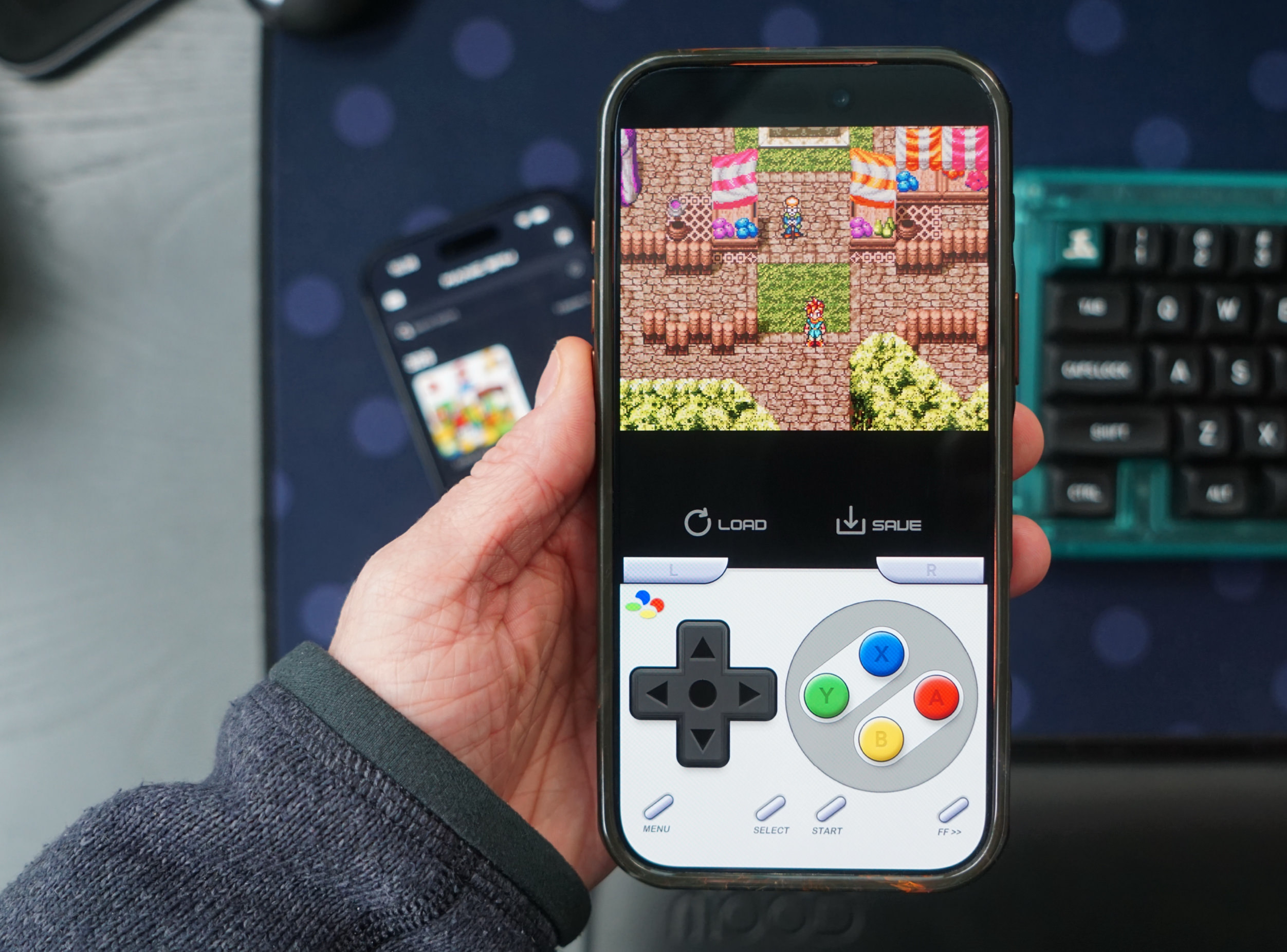 Eine Hand, die ein Smartphone hält, auf dem ein Retro-RPG-Videospiel mit einem virtuellen Gamepad überlagert angezeigt wird; ein zweites Telefon, eine Tastatur und eine Maus sind im Hintergrund sichtbar.