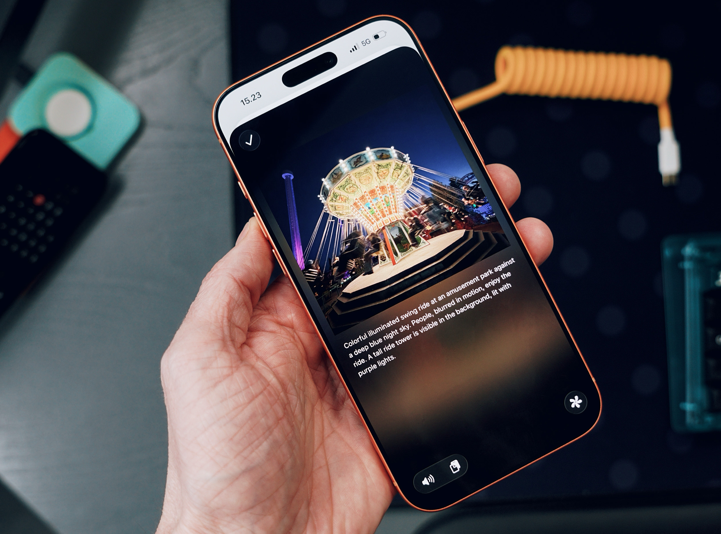 Hand hält ein Telefon, das ein Foto eines hell erleuchteten Kettenkarussells in einem Vergnügungspark vor einem tiefblauen Nachthimmel zeigt, mit verschwommenen Menschen in Bewegung und einem hohen, violett beleuchteten Turm im Hintergrund.