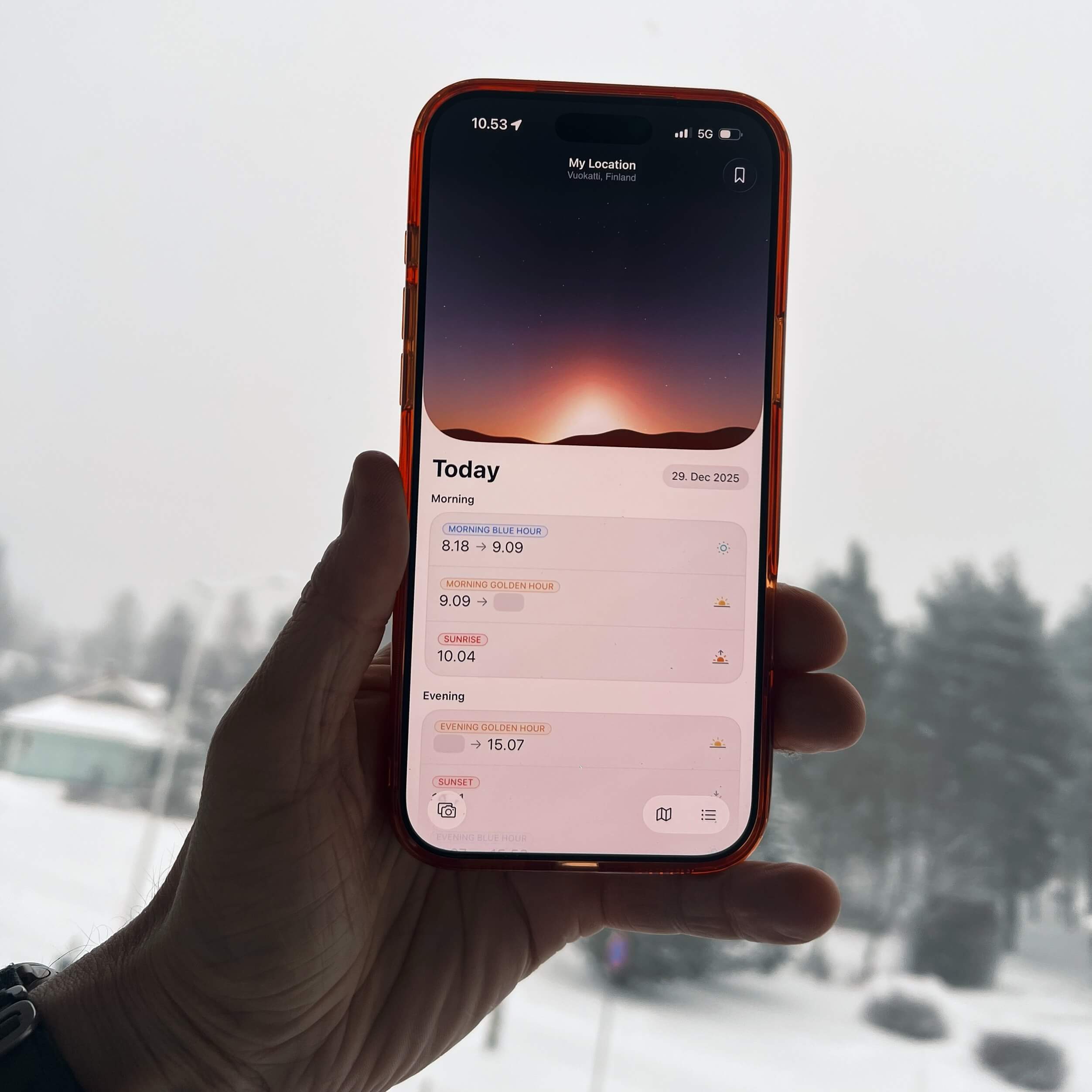 Eine Hand, die ein rot gerahmtes Smartphone hält, das eine "Heute"-Sonnenereignis-App vor einem verschneiten Hintergrund im Freien anzeigt. Die App zeigt verschiedene Sonnenauf- und -untergangszeiten für den 29. Dezember 2025 an.
