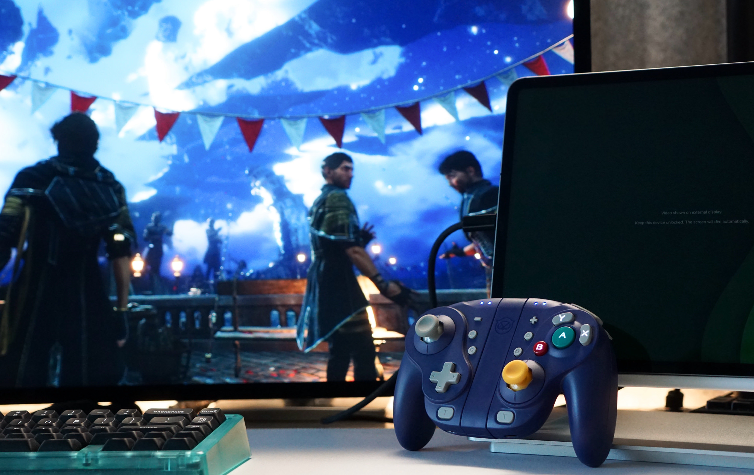 A purple GameCube controller is positioned in front of a monitor displaying a fantasy game scene. The scene includes characters in a medieval setting, beneath a starry sky with decorative pennants. A laptop screen is to the right with video displayed on an external monitor and a backlit keyboard is below the monitor.
