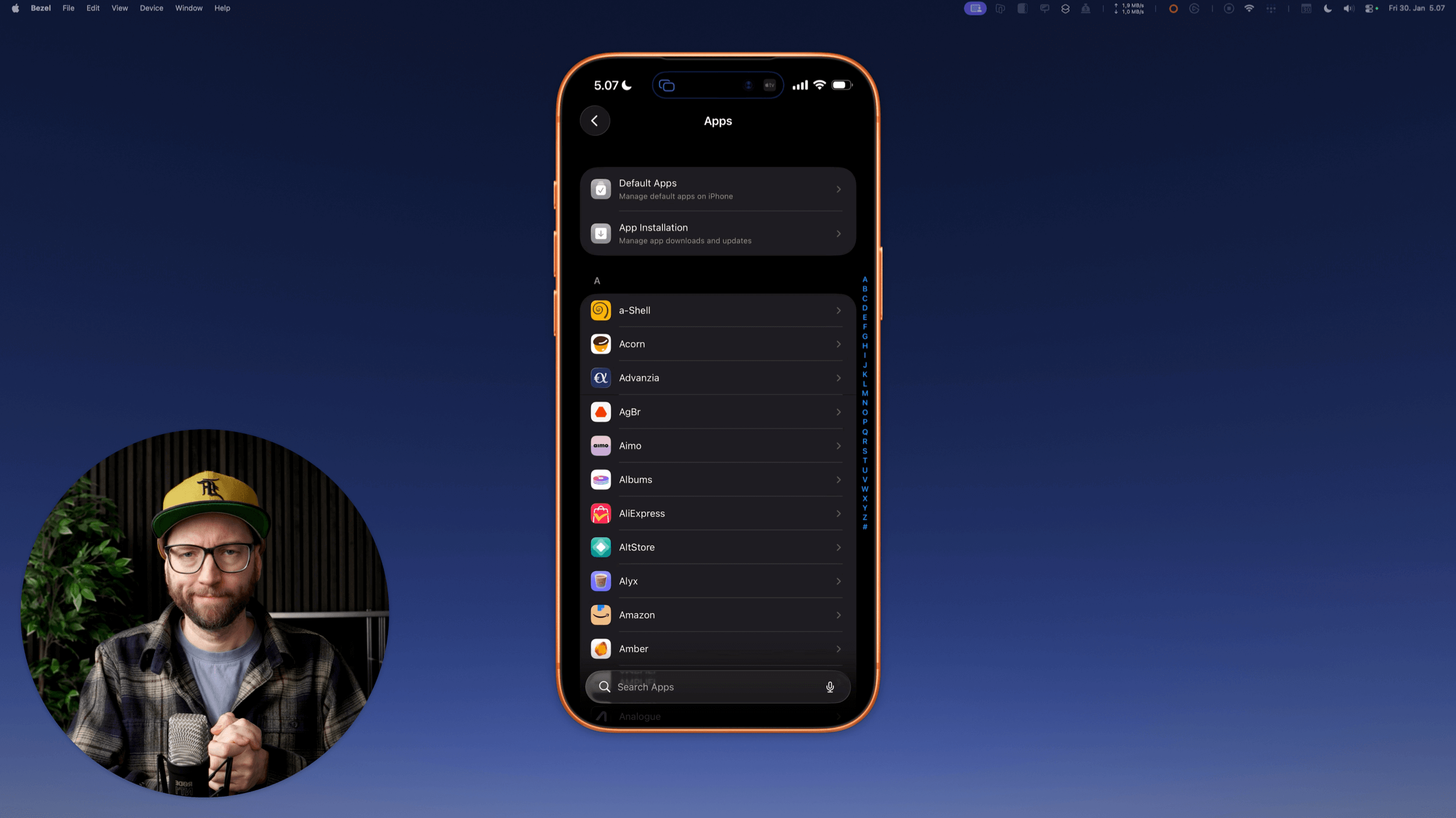 Ein Mann mit Brille und Baseballkappe ist vor einem blauen Hintergrund abgebildet, der ein offenes iPhone mit dem Einstellungsbildschirm "Apps" zeigt. Das iPhone zeigt eine Liste von Apps mit Optionen wie "Standard-Apps" und "App-Installation". Der Mann befindet sich in einem Studio, hält ein Mikrofon.