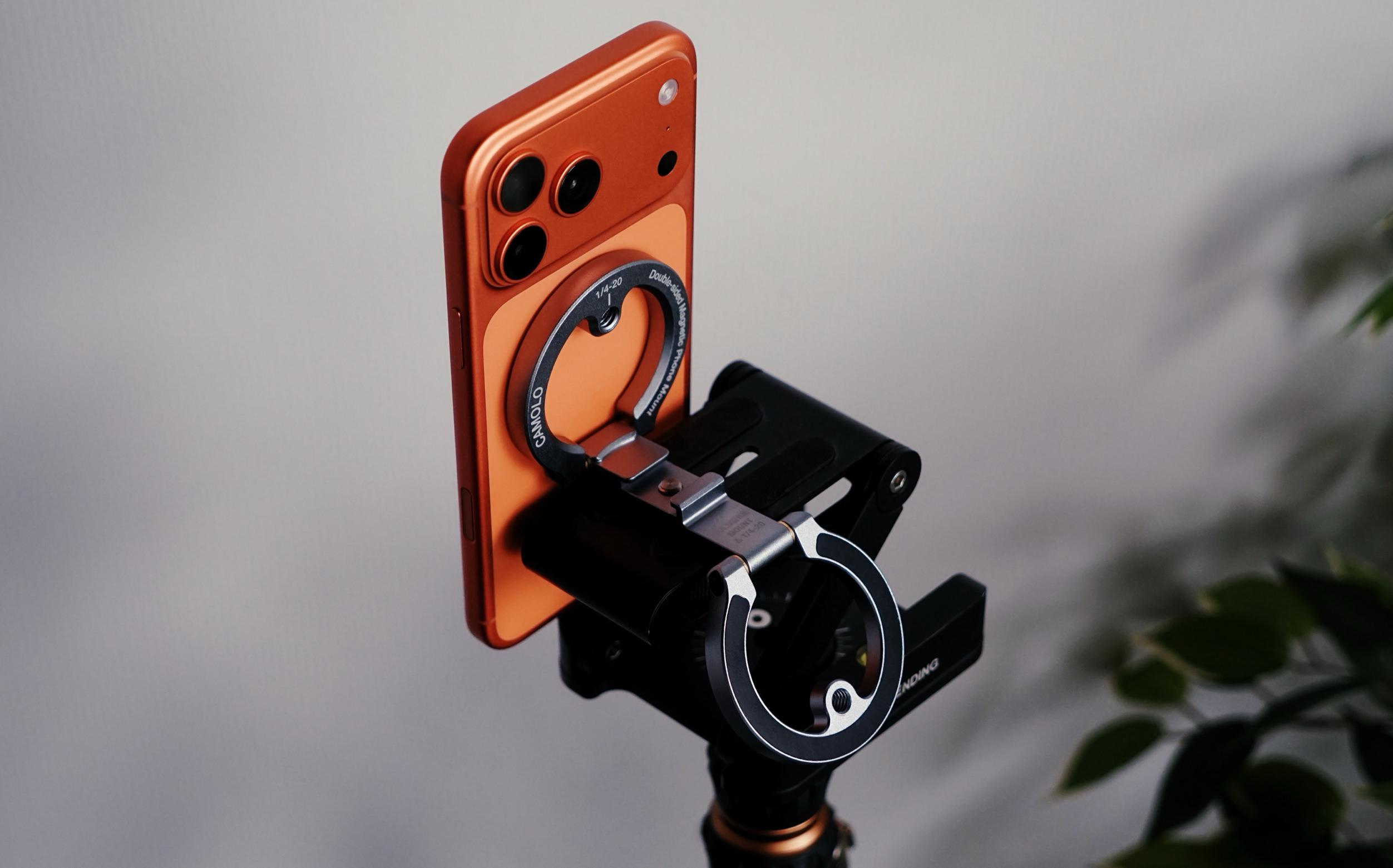 Ein orangefarbenes Smartphone mit einem Ringgriff, der magnetisch an einer schwarzen Telefonhalterung befestigt ist, die auf einem Stativ positioniert ist.
