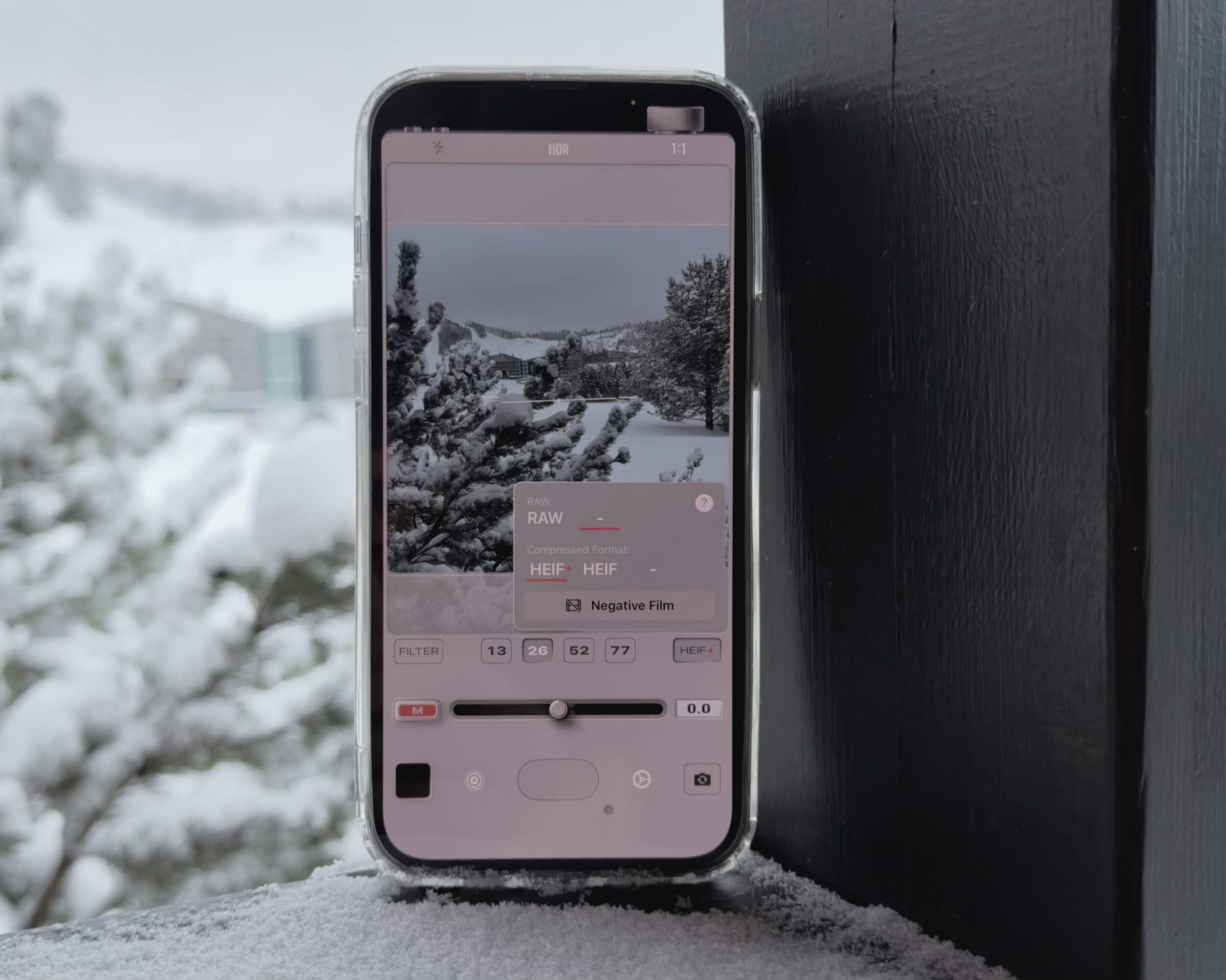 Ein Smartphone, das eine Kamera-App mit einer verschneiten Landschaft im Sucher anzeigt, wird gegen eine schneebedeckte Oberfläche und eine schwarz lackierte Holzkonstruktion gehalten.