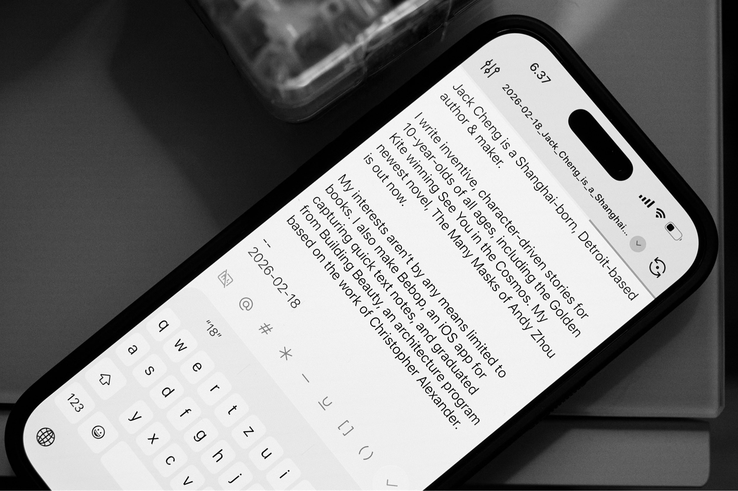 Ein Graustufenbild zeigt ein Smartphone, das Text anzeigt. Der Text in weißer Schrift auf hellem Hintergrund wird auf einem schwarzen Bildschirm angezeigt, wobei die Tastatur unten sichtbar ist. Die Zeit "6:37" steht oben rechts, gefolgt von: "Jack Cheng ist ein in Shanghai geborener, in Detroit ansässiger Autor und Macher. Ich schreibe einfallsreiche, von Charakteren angetriebene Geschichten für 10-Jährige jeden Alters, darunter die Golden Kite, die "See You in the Cosmos" gewonnen hat. Mein neuester Roman, "The Many Masks of Andy Zhou", ist jetzt erhältlich. Meine Interessen beschränken sich keineswegs auf Bücher. Ich mache auch Bebop, eine iOS-App zum Erfassen von Schnelltextnotizen, und habe das Architekturprogramm Building Beauty auf der Grundlage der Arbeit von Christopher Alexander absolviert."
