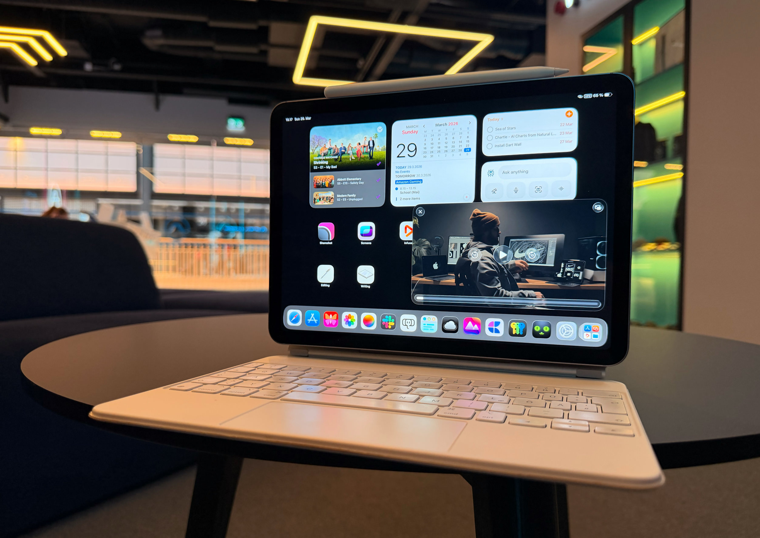 Ein Tablet mit angeschlossener Tastatur steht auf einem runden schwarzen Tisch in einer hell beleuchteten, modernen Bürolounge. Der Bildschirm zeigt eine iPadOS-Oberfläche mit Widgets für einen Kalender und eine Aufgabenliste, App-Symbole und ein schwebendes Videoplayer-Fenster, in dem eine Person vor Computerbildschirmen zu sehen ist.