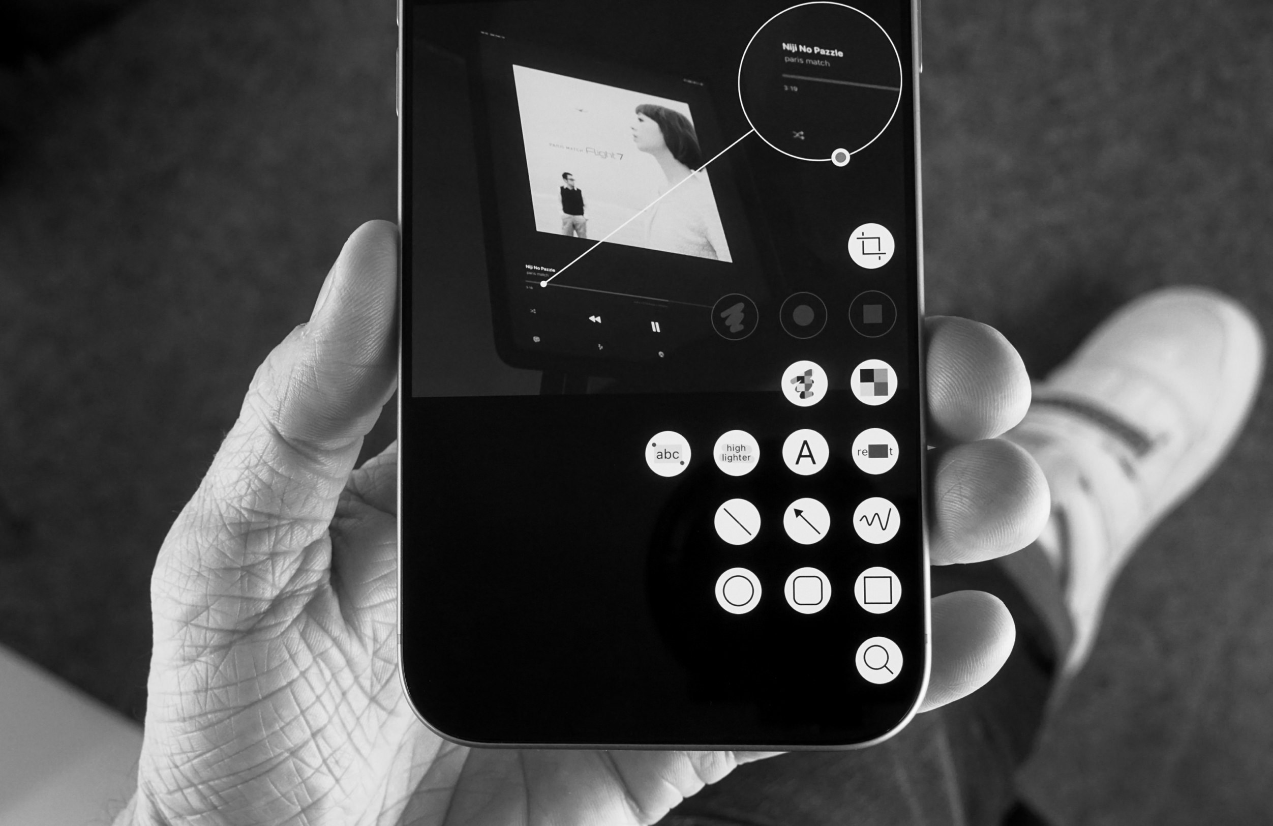 Eine Hand hält ein Smartphone, das eine Musikwiedergabeoberfläche mit verschiedenen Symbolen anzeigt. Auf dem Bildschirm steht: "Nip No Puzzle" und "paris match".