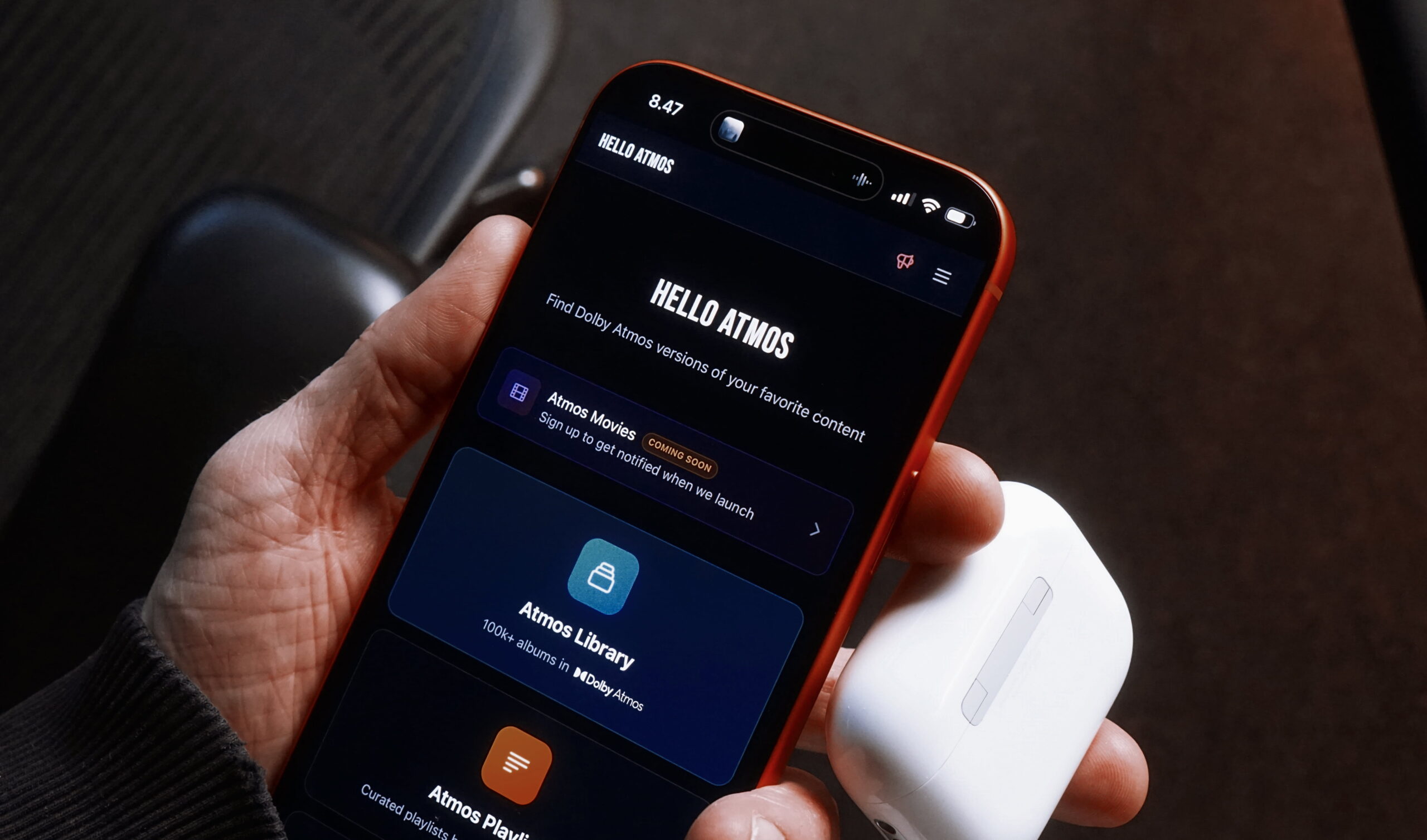 Eine Hand, die ein rotes iPhone hält, das die "Hello Atmos"-App anzeigt, mit einem AirPods-Etui in derselben Hand; die App-Oberfläche zeigt Optionen zum Finden von Dolby Atmos-Versionen von Inhalten, einschließlich Atmos Movies und Atmos Library mit über 100.000 Alben.

