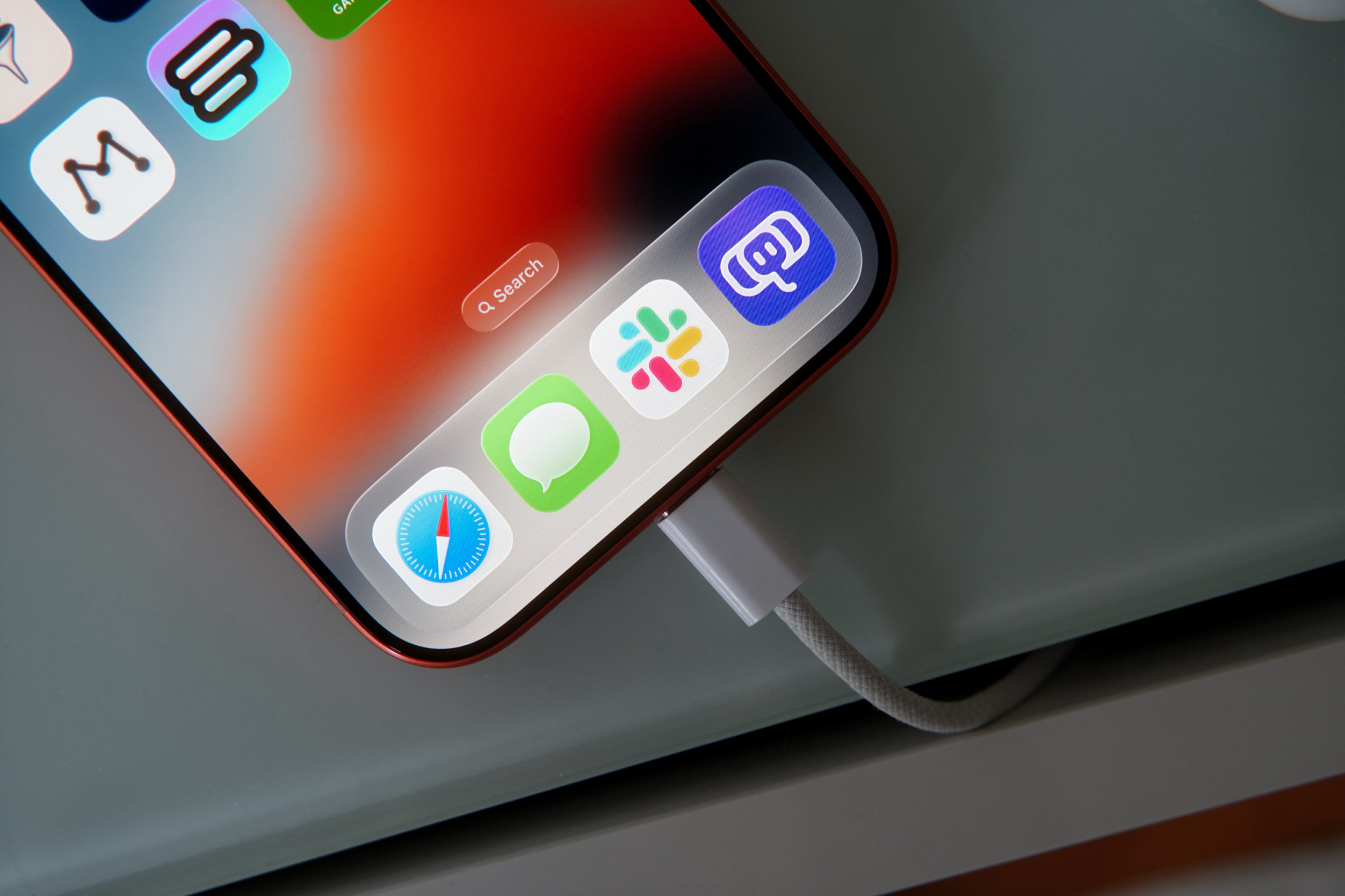 Ein Smartphone mit einem farbenfrohen Bildschirm, auf dem diverse App-Symbole zu sehen sind, wird mit einem weißen Ladekabel auf einer hellen Oberfläche geladen. Auf dem Bildschirm befindet sich eine Suchleiste mit dem Text „Search“.