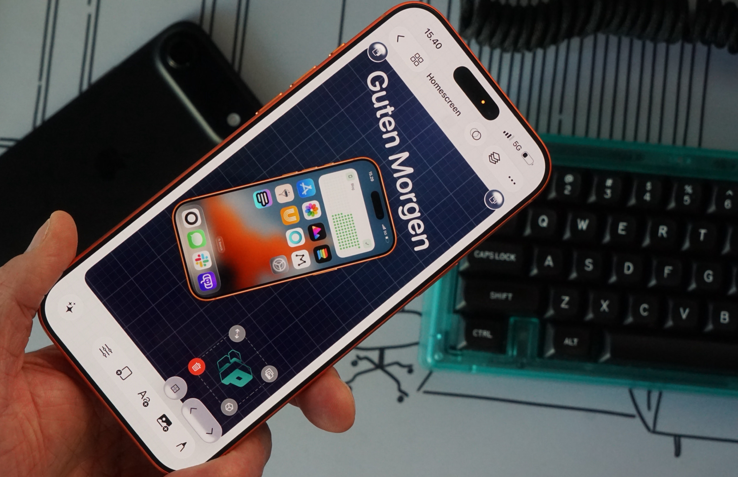 Eine Hand hält ein Smartphone mit orangefarbenem Gehäuserand, auf dessen Display eine App mit einem Gitter-Hintergrund und einer Vorschau eines iPhone-Startbildschirms mit verschiedenen App-Symbolen zu sehen ist. Auf dem Bildschirm sind die Texte „Guten Morgen“, „15.40“ und „Homescreen“ lesbar. Im Hintergrund liegen ein zweites, dunkles Smartphone und eine türkisfarbene mechanische Tastatur auf einem Schreibtisch.