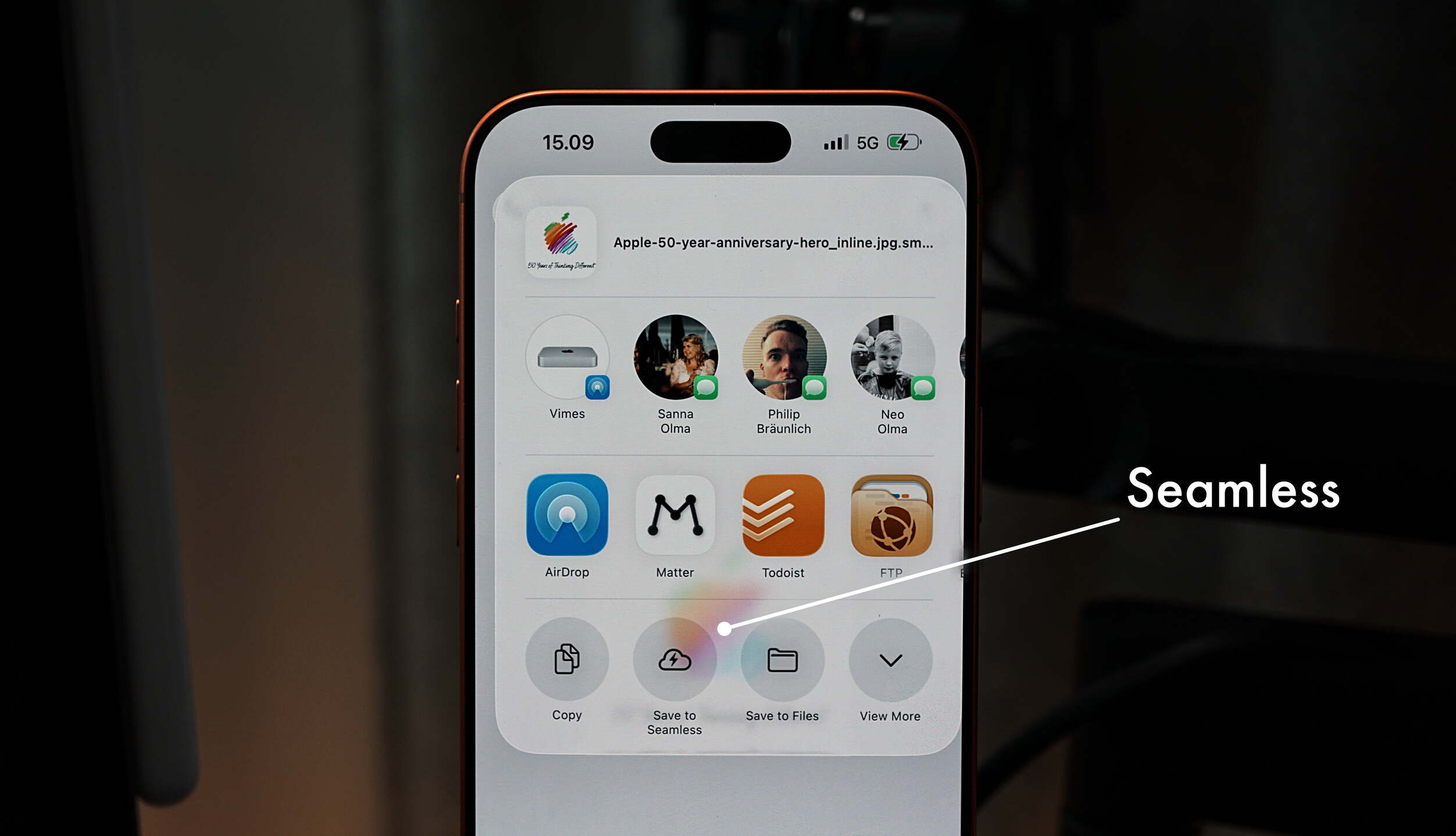 Eine Nahaufnahme des Bildschirms eines iPhones, das das Freigabeblatt anzeigt. Der obere Bildschirmrand zeigt die Uhrzeit 15.09 und ein 5G-Symbol an. Das Freigabeblatt zeigt App-Symbole für "Vimes", "Sanna Olma", "Philip Bräunlich", "Neo Olma", "AirDrop", "Matter", "Todoist" und "FTP" an, wobei "Save to Seamless" hervorgehoben ist. Ein Pfeil zeigt vom Wort "Seamless" auf das entsprechende Symbol. Das Apple-Logo ist ebenfalls sichtbar. Der Text "50 Years of Thinking Different" und der Dateiname "Apple-50-year-anniversary-hero_inline.jpg.sm..." sind ebenfalls sichtbar.