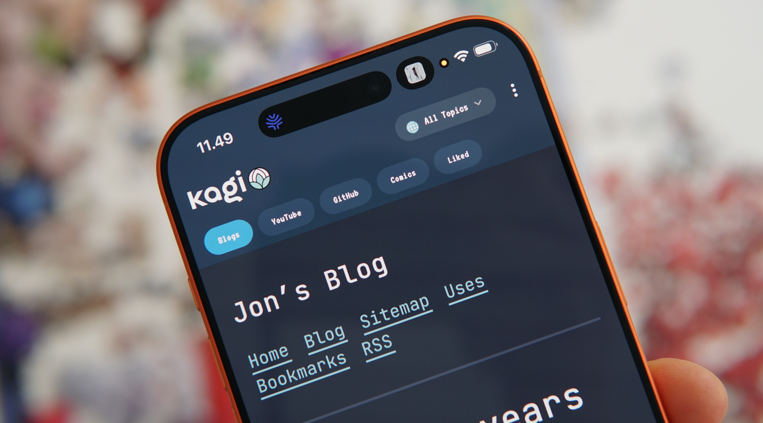 Eine Hand, die ein orangefarbenes Telefon hält, auf dem eine Blog-Website angezeigt wird. Die Website trägt den Titel "Jon's Blog" und enthält Navigationslinks wie "Home", "Blog", "Sitemap", "Uses", "Bookmarks" und "RSS". Der Header der Website enthält das Logo "kagi" und Schaltflächen für "Blogs", "YouTube", "GitHub", "Comics" und "Liked". Die Oberseite des Telefons zeigt die Uhrzeit "11.49" und einen Kameraausschnitt.
