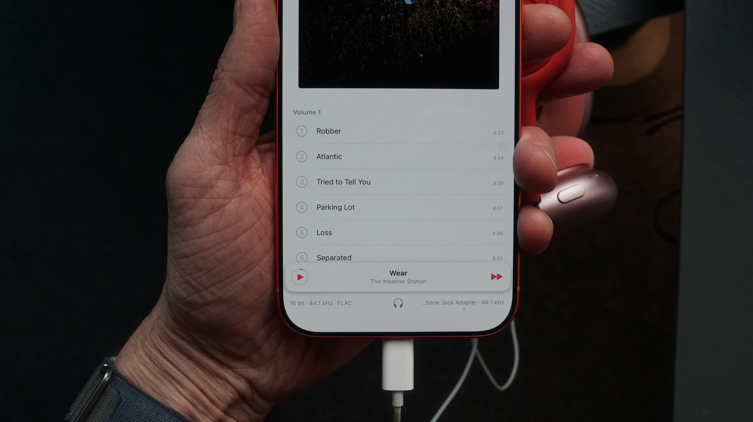 Eine Hand hält ein rotes Smartphone, auf dem eine Musik-App-Oberfläche angezeigt wird. Der Bildschirm zeigt eine Playlist mit dem Titel "Volume 1" mit einer Liste von Songs: "Robber", "Atlantic", "Tried to Tell You", "Parking Lot", "Loss" und "Separated". Der aktuell gespielte Song ist "Wear" von "The Weather Station". Das Telefon ist mit einem weißen Kopfhöreradapter verbunden.
