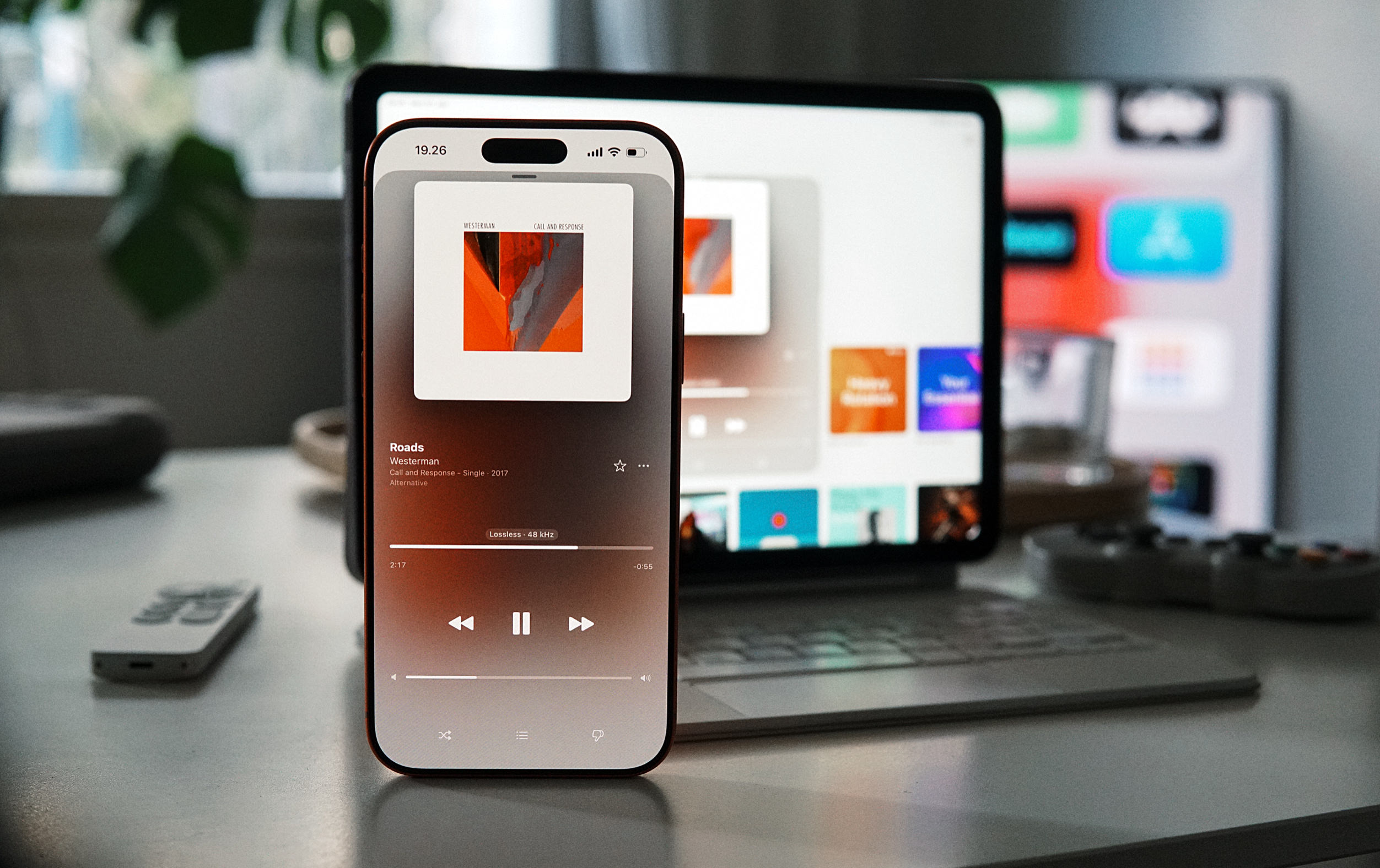 Eine Nahaufnahme eines Smartphones auf einem Schreibtisch, das die Benutzeroberfläche einer Musik-Streaming-App mit Albumcover und Wiedergabesteuerung anzeigt. Im Hintergrund steht ein Tablet mit sichtbaren App-Symbolen auf dem Bildschirm.