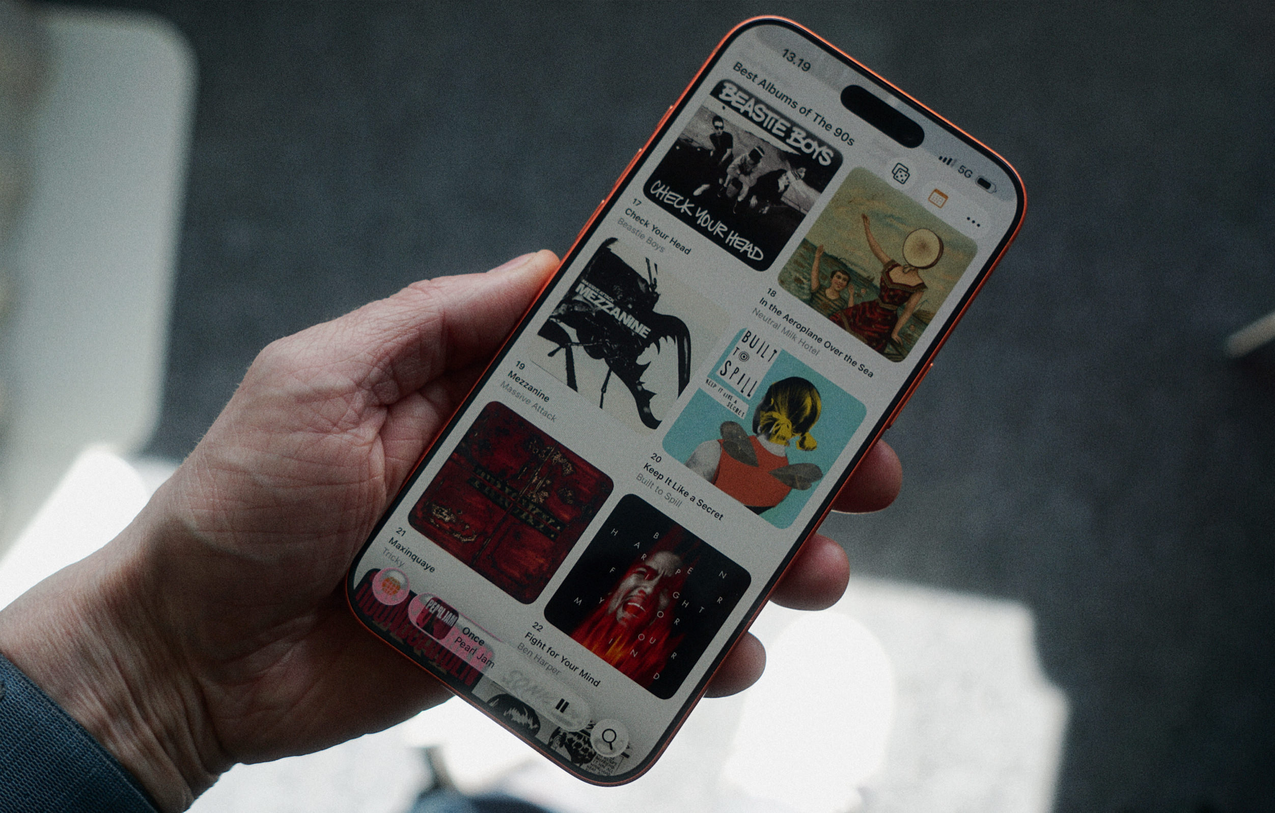 Eine Hand hält ein orangefarbenes Smartphone, auf dessen Display eine Liste mit dem Titel „Best Albums of The 90s“ zu sehen ist. Die App zeigt verschiedene Albumcover und folgende Texte: „13.19“, „Best Albums of The 90s“, „17 Check Your Head Beastie Boys“, „18 In the Aeroplane Over the Sea Neutral Milk Hotel“, „19 Mezzanine Massive Attack“, „20 Keep It Like a Secret Built to Spill“, „21 Maxinquaye Tricky“, „22 Fight for Your Mind Ben Harper“ sowie am unteren Rand einen Musik-Player mit der Anzeige „Once Pearl Jam“.