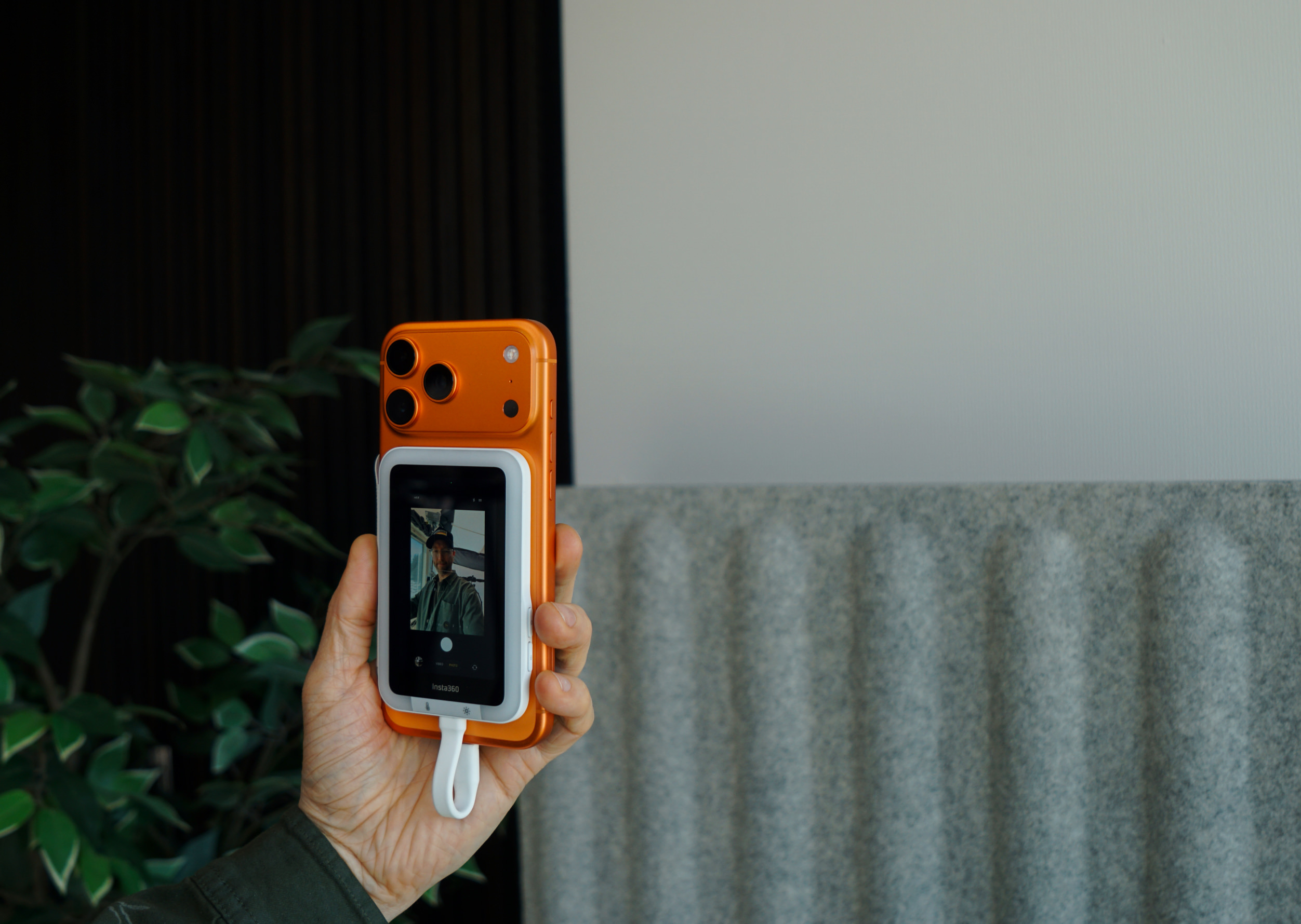 Eine Hand hält ein leuchtend oranges Smartphone mit einem weißen Aufsatz auf der Rückseite. Das weiße Gerät verfügt über ein kleines digitales Display, das eine Live-Kameraaufnahme einer Person mit Kappe und Jacke zeigt. Der Text „Insta360“ ist am unteren Rand des Bildschirms des Aufsatzes deutlich zu erkennen. Der Hintergrund umfasst eine Zimmerpflanze und eine graue, strukturierte Wand.