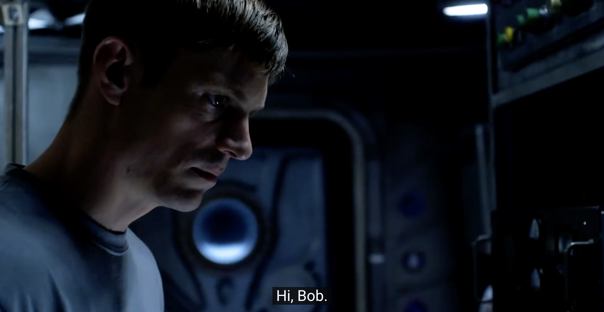 Ein Mann mit kurzen Haaren und ernstem Gesichtsausdruck blickt in einem schwach beleuchteten, industriell wirkenden Raum nachdenklich auf etwas außerhalb des Bildschirms. Der Untertitel lautet: „Hi, Bob.“