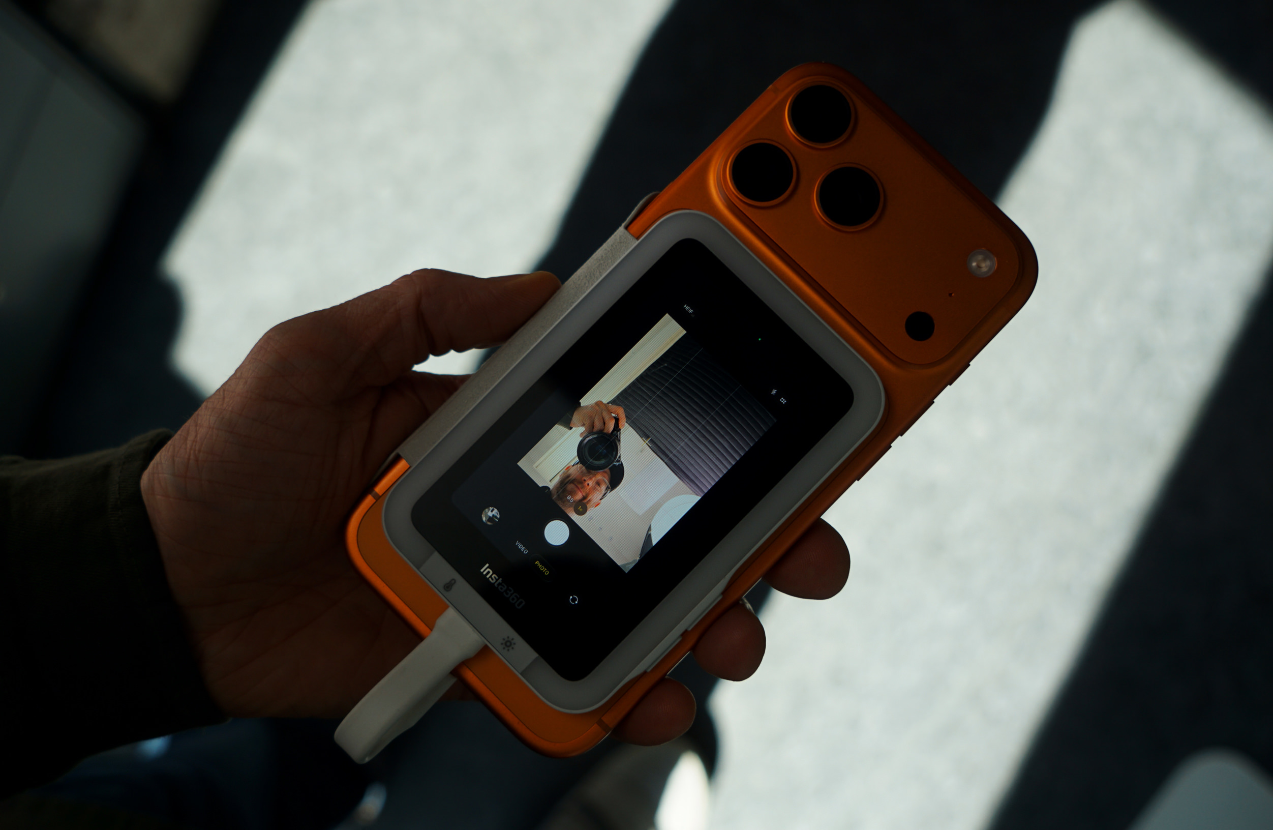 Eine Hand hält ein orangefarbenes Kamerazubehör, das an einem Smartphone befestigt ist. Auf dem Bildschirm ist eine Live-Kameraansicht zu sehen, die die Person beim Aufnehmen des Bildes zeigt. Am unteren Bildschirmrand steht der Text „Insta360“.