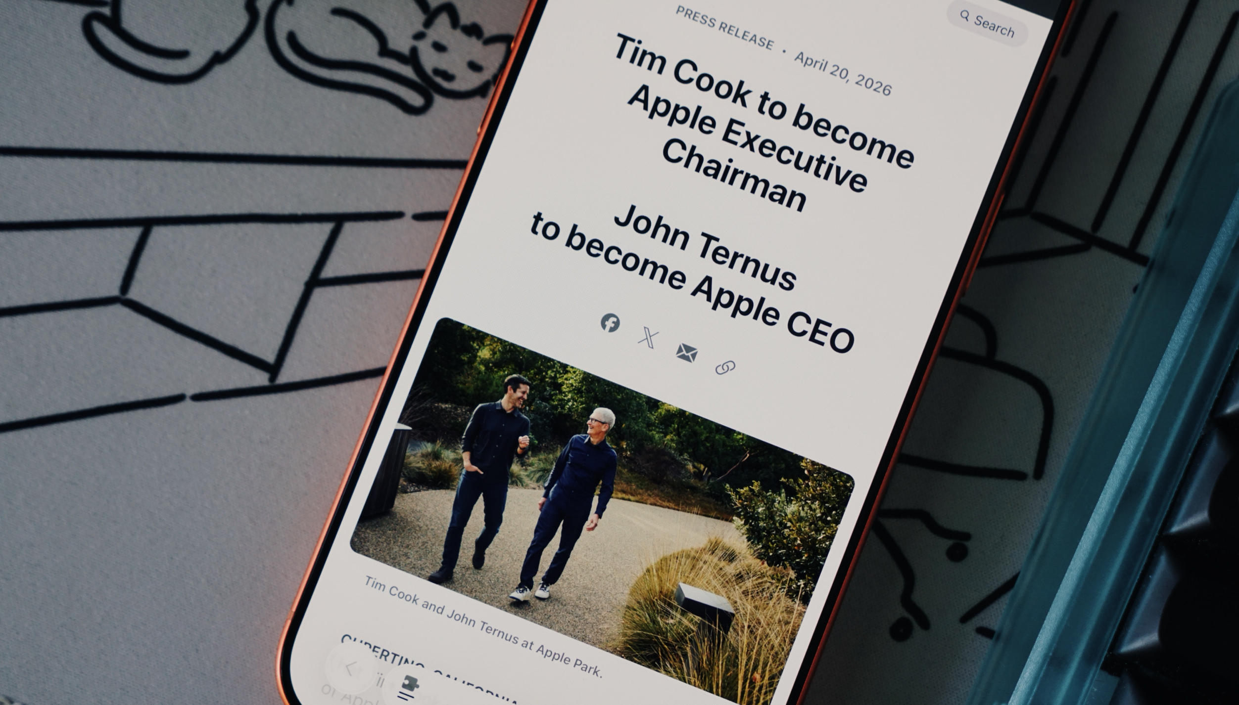 Ein Smartphone zeigt eine digitale Pressemitteilung über Führungswechsel bei einem Technologieunternehmen. Ein beigefügtes Foto zeigt zwei Männer, die auf einem befestigten Weg im Freien spazieren. Der Text auf dem Bildschirm lautet: PRESS RELEASE, April 20, 2026 Tim Cook to become Apple Executive Chairman John Ternus to become Apple CEO Tim Cook and John Ternus at Apple Park. CUPERTINO CALIFORNIA"