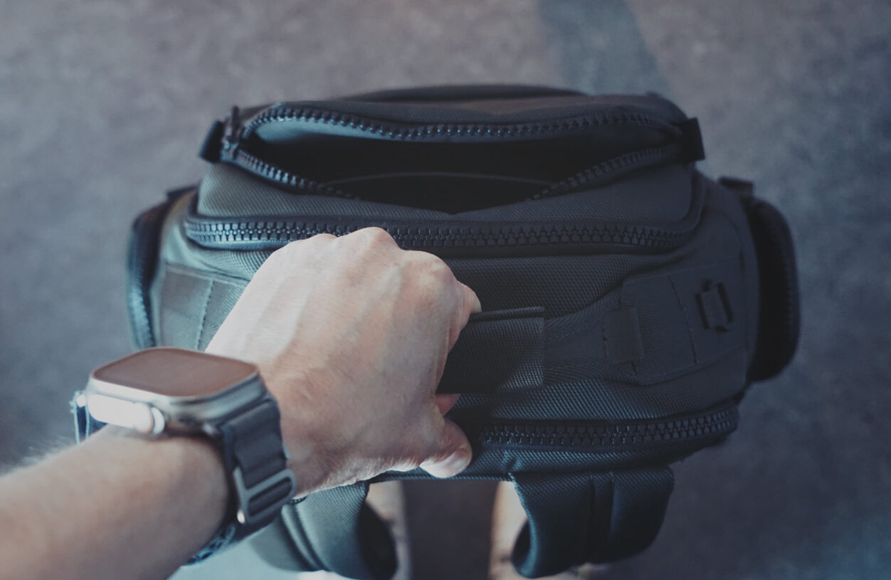 Eine Hand mit einer Smartwatch hält den oberen Tragegriff eines dunklen Rucksacks.