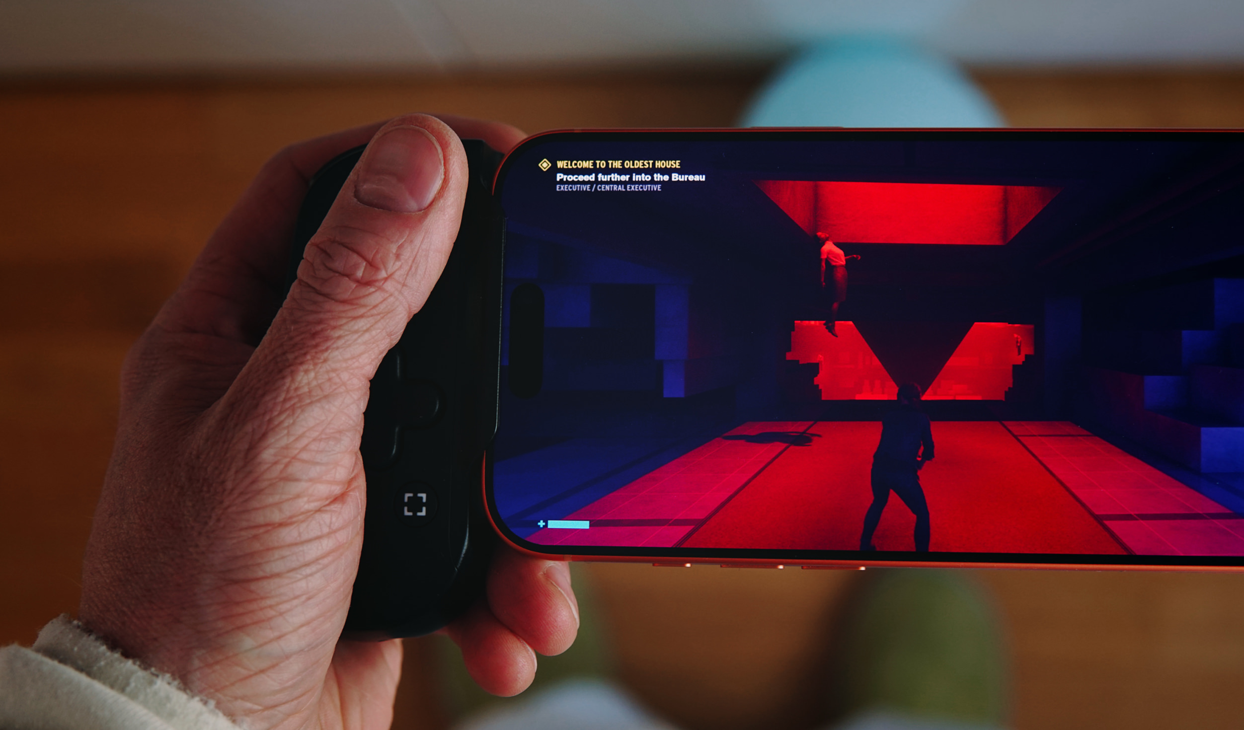 Eine Nahaufnahme einer Hand, die ein mit einem Spiele-Controller ausgestattetes Smartphone hält, das eine Videospielszene in einem düsteren, rot beleuchteten Raum mit einer schwebenden Figur zeigt.