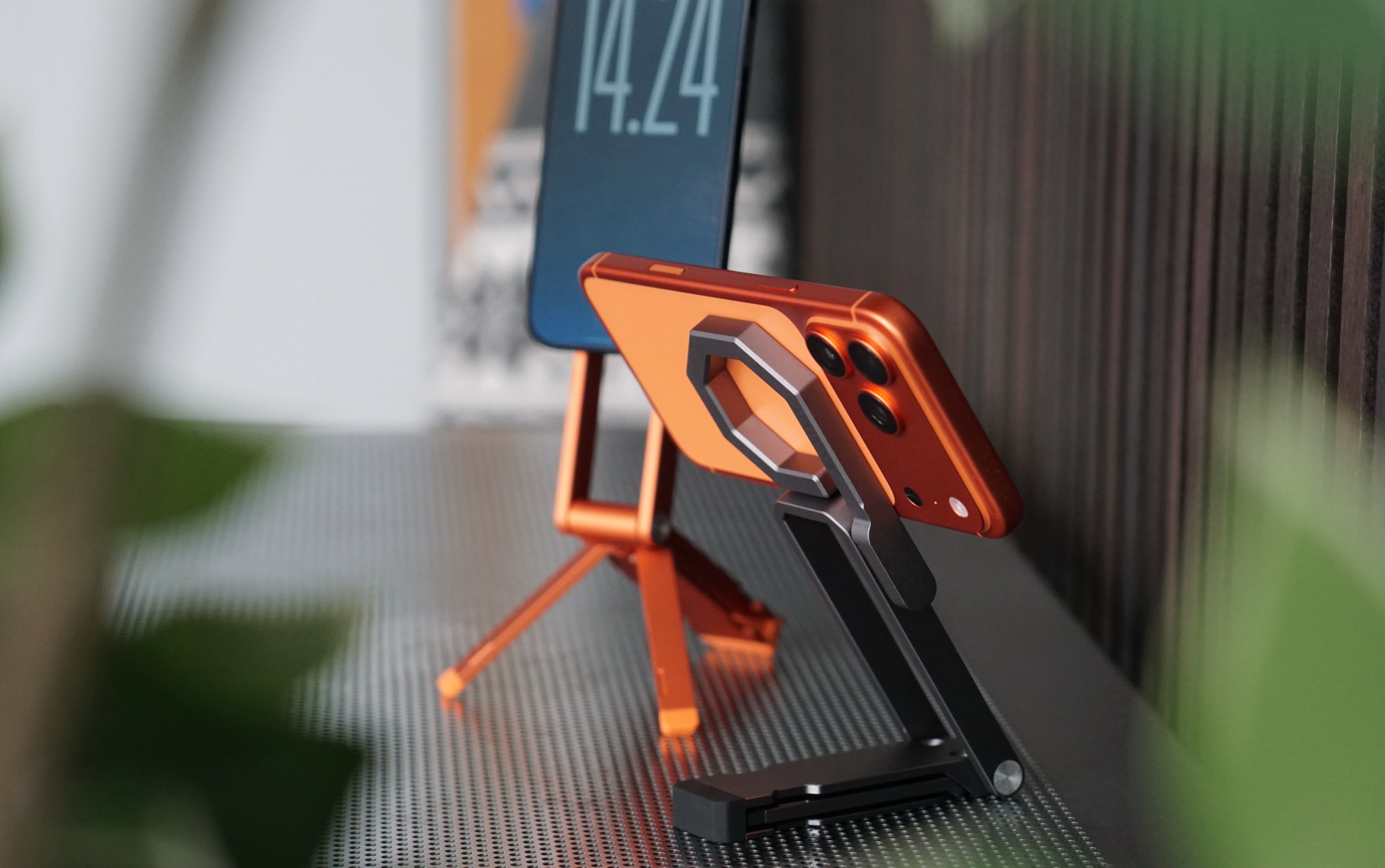 Ein oranges Smartphone steht in einem schwarzen, klappbaren Halter im Vordergrund, während ein weiteres Smartphone auf einem orangefarbenen Stativ im Hintergrund platziert ist und auf seinem Bildschirm die Uhrzeit „14.24“ anzeigt. Die Geräte befinden sich auf einer metallischen, perforierten Oberfläche.