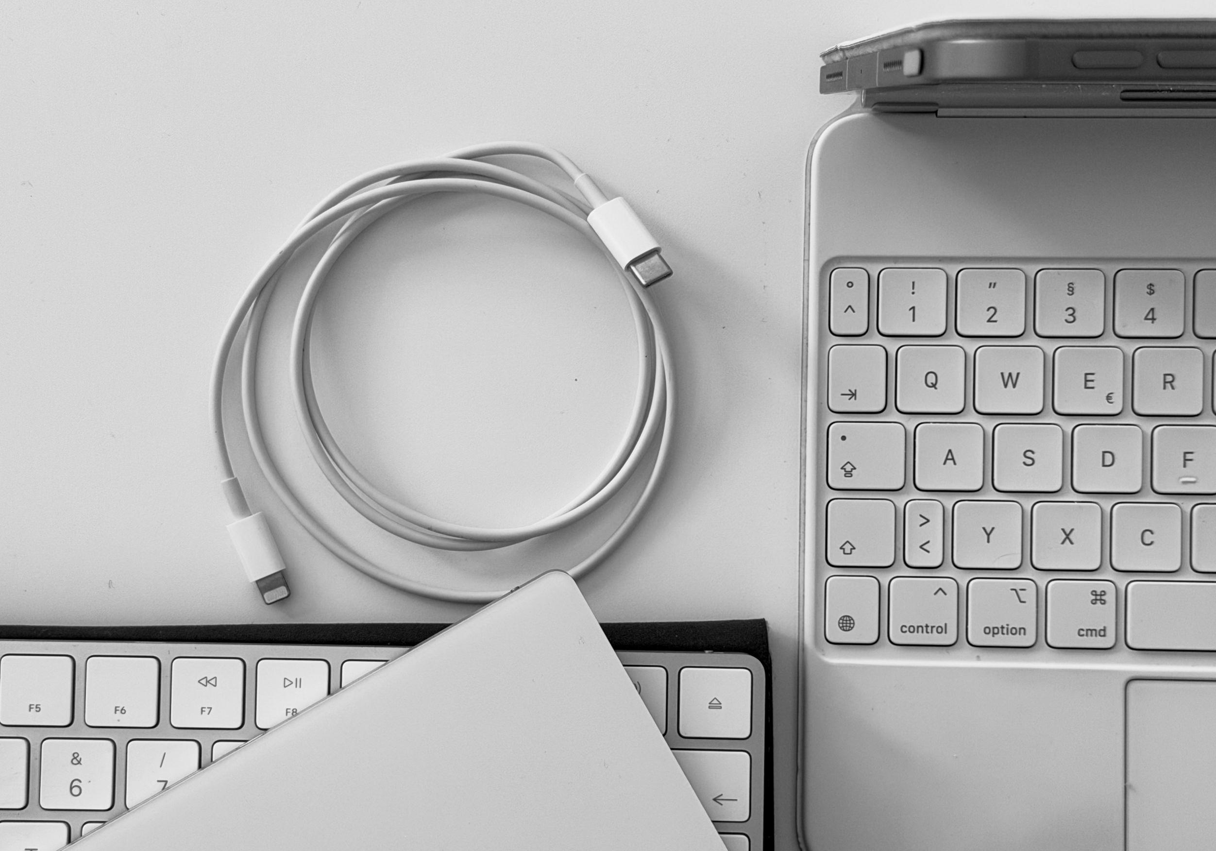 Eine Schwarz-Weiß-Ansicht von oben auf einen Arbeitsbereich, die ein weißes, aufgerolltes Ladekabel zwischen einer externen Tastatur und einem Tablet mit integrierter Tastatur zeigt.