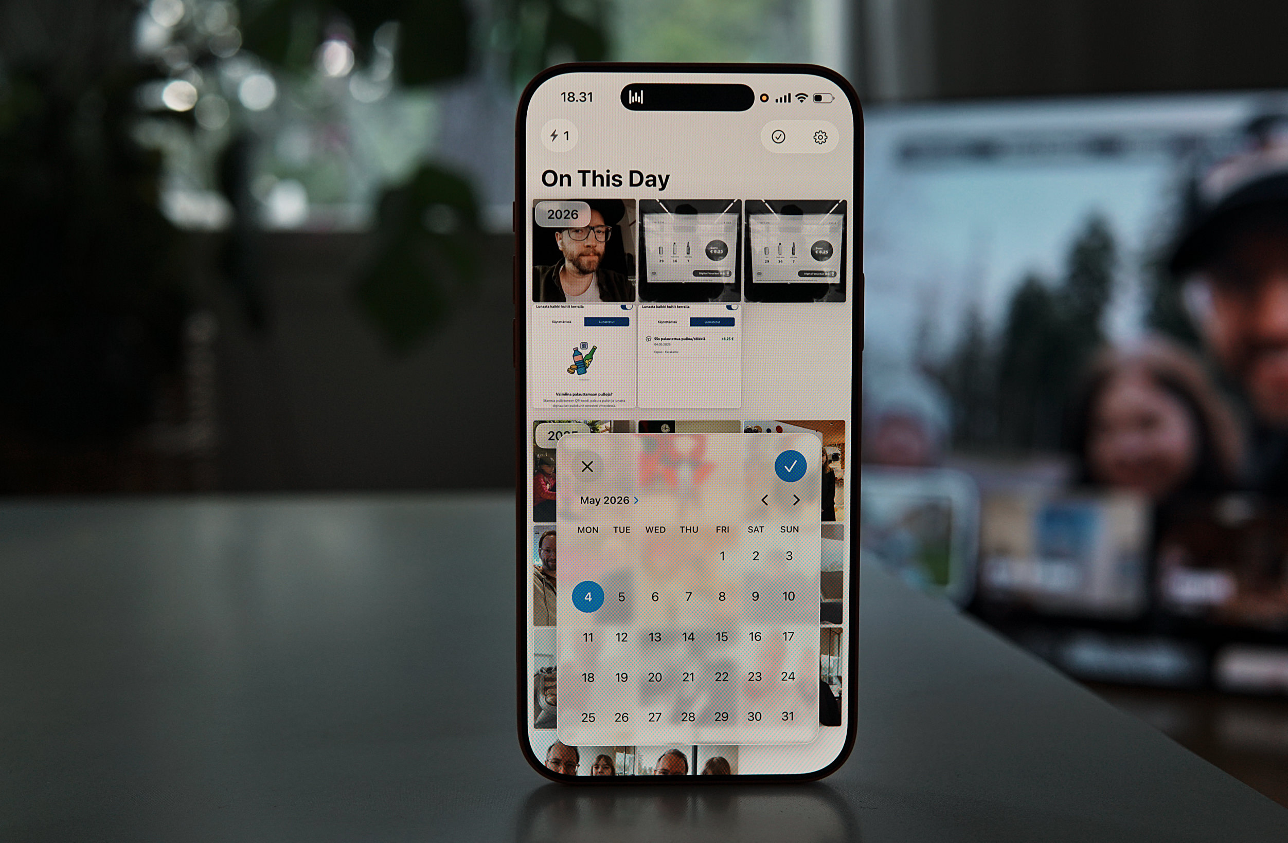 Ein Smartphone zeigt eine Fotogalerie-App mit der Überschrift „On This Day“. Ein halbtransparentes Kalenderfenster ist geöffnet, das den „Mai 2026“ anzeigt, wobei der 4. Tag blau markiert ist. Im Hintergrund sind unscharfe Bildvorschauen zu sehen.