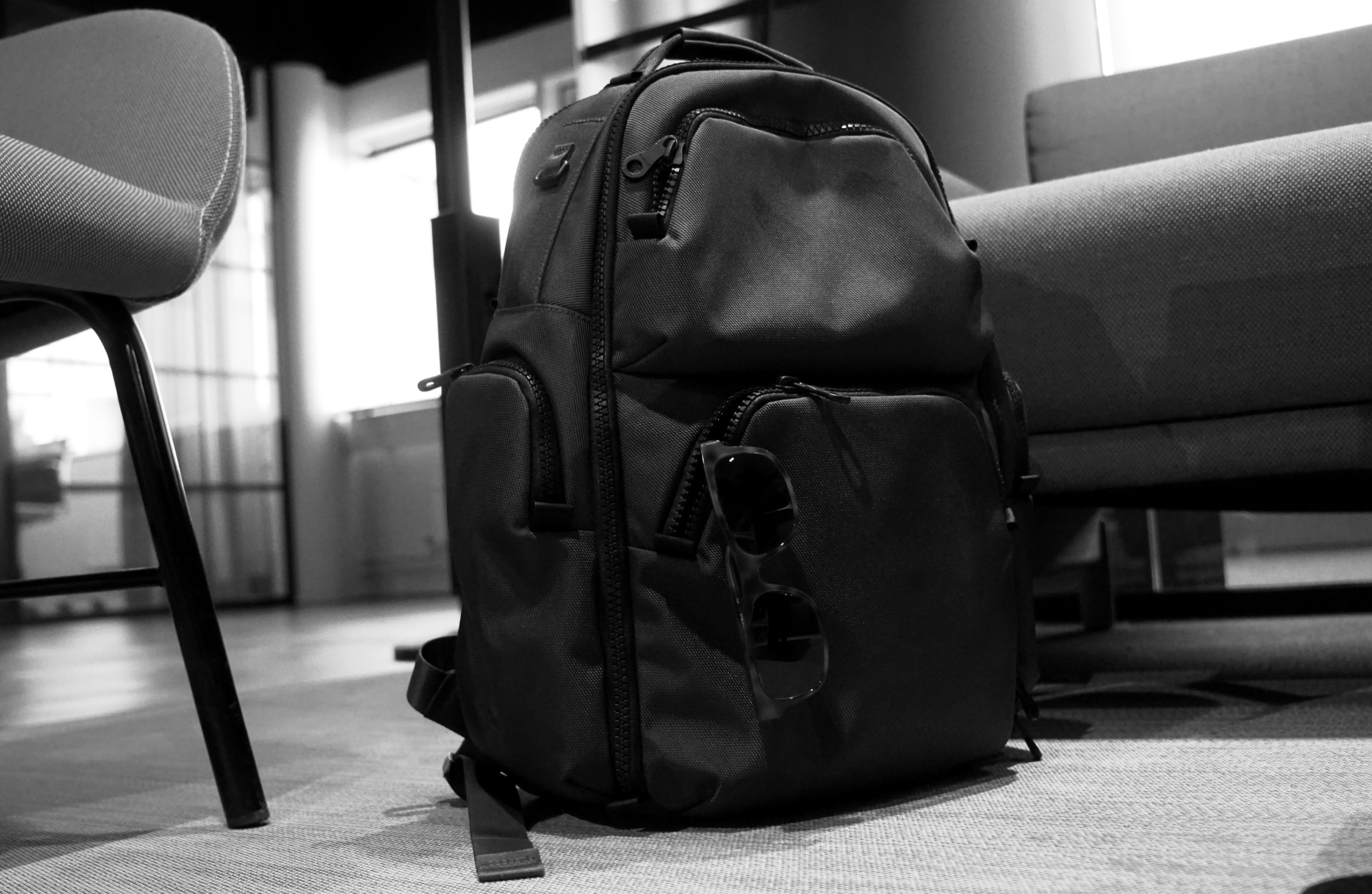 Ein schwarzer Rucksack steht auf dem Boden in einem büroähnlichen Raum zwischen einem Stuhl und einem Sofa, wobei eine Sonnenbrille am vorderen Reißverschluss befestigt ist.