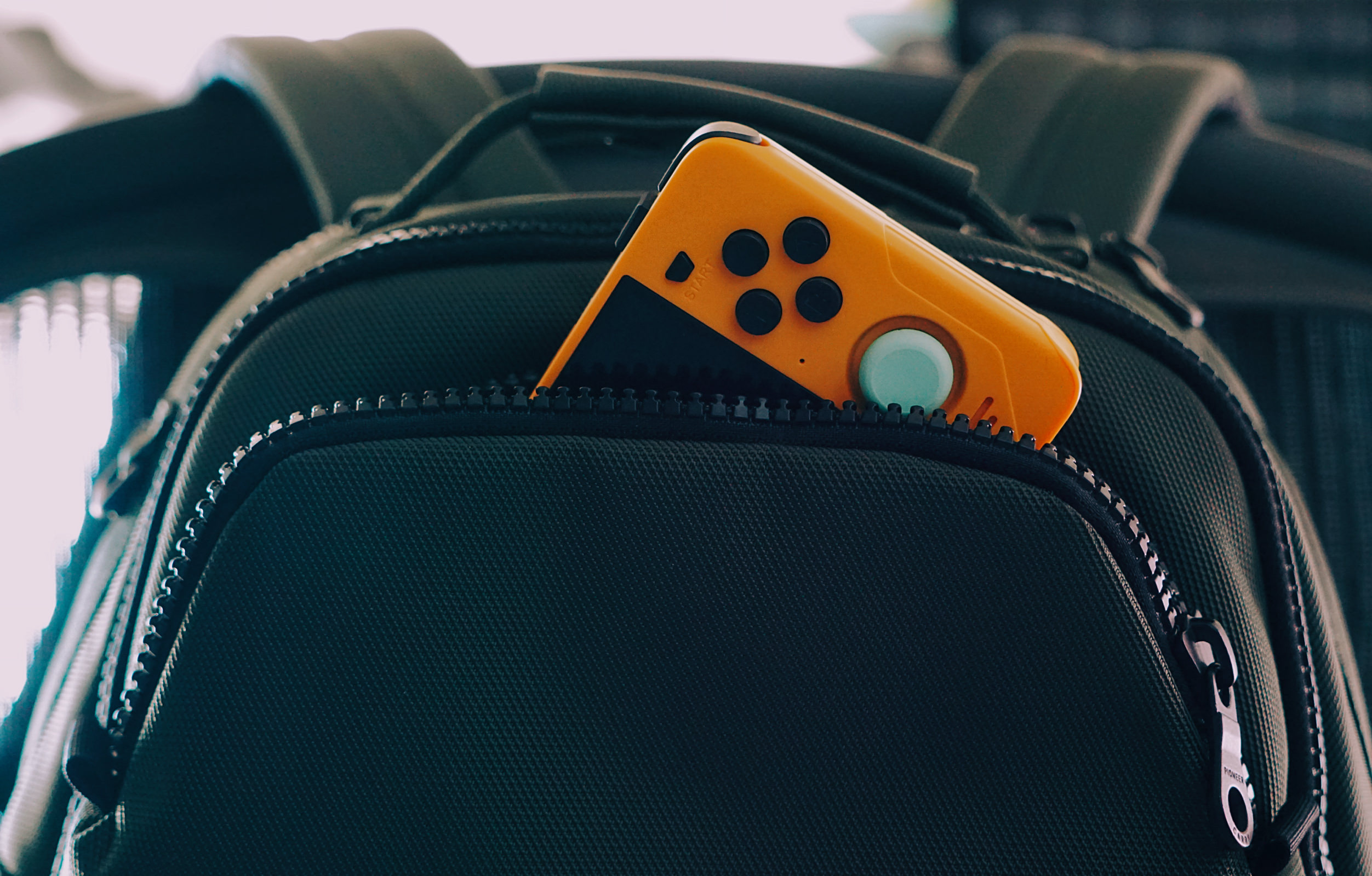 Eine kleine, gelbe, tragbare Spielkonsole mit einer markanten Kurbel und schwarzen Tasten, die halb in der Vordertasche eines dunklen Rucksacks steckt.
