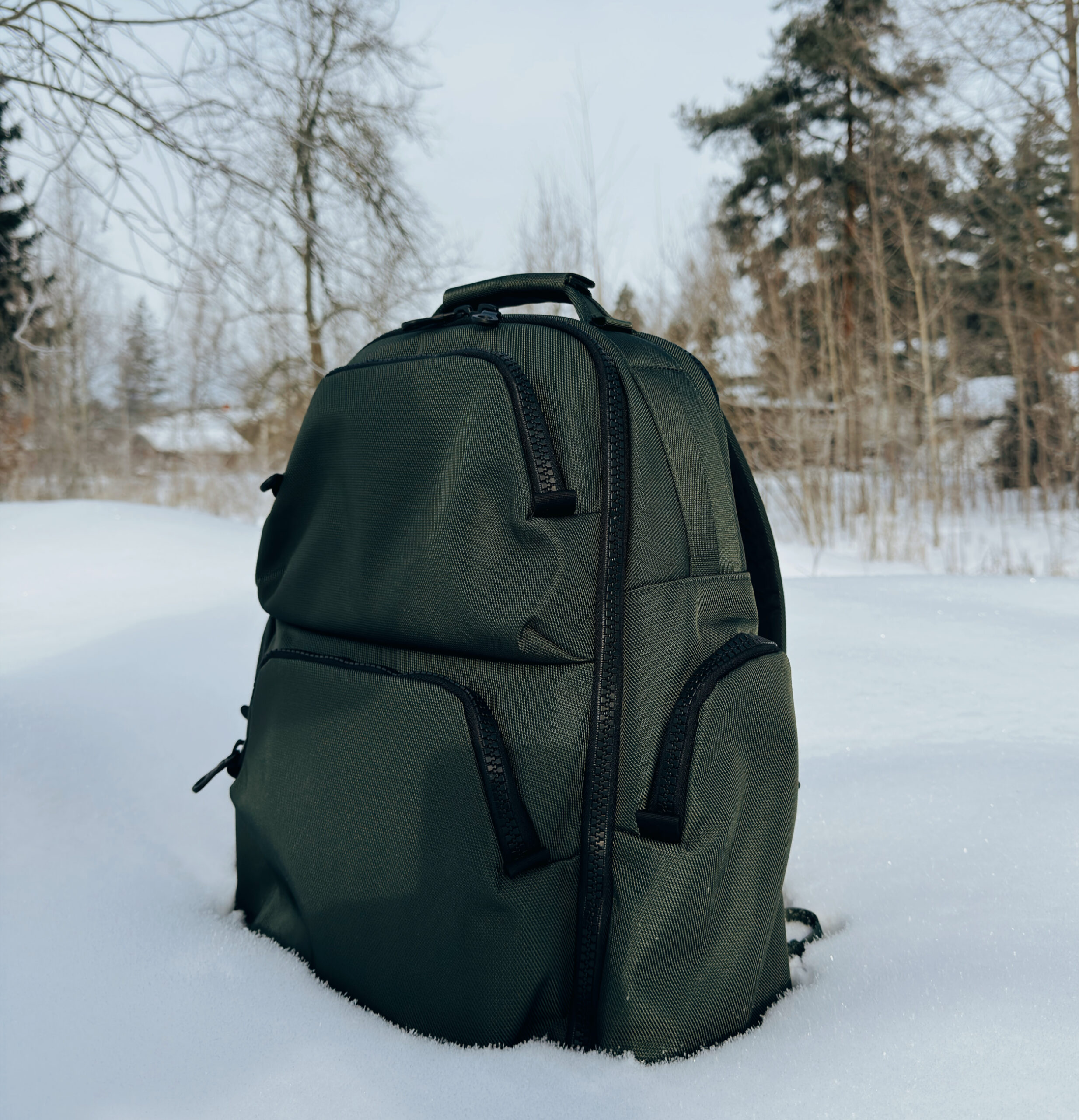 Ein dunkelgrüner Rucksack steht aufrecht im tiefen, frischen Schnee vor einem unscharfen Winterwald.