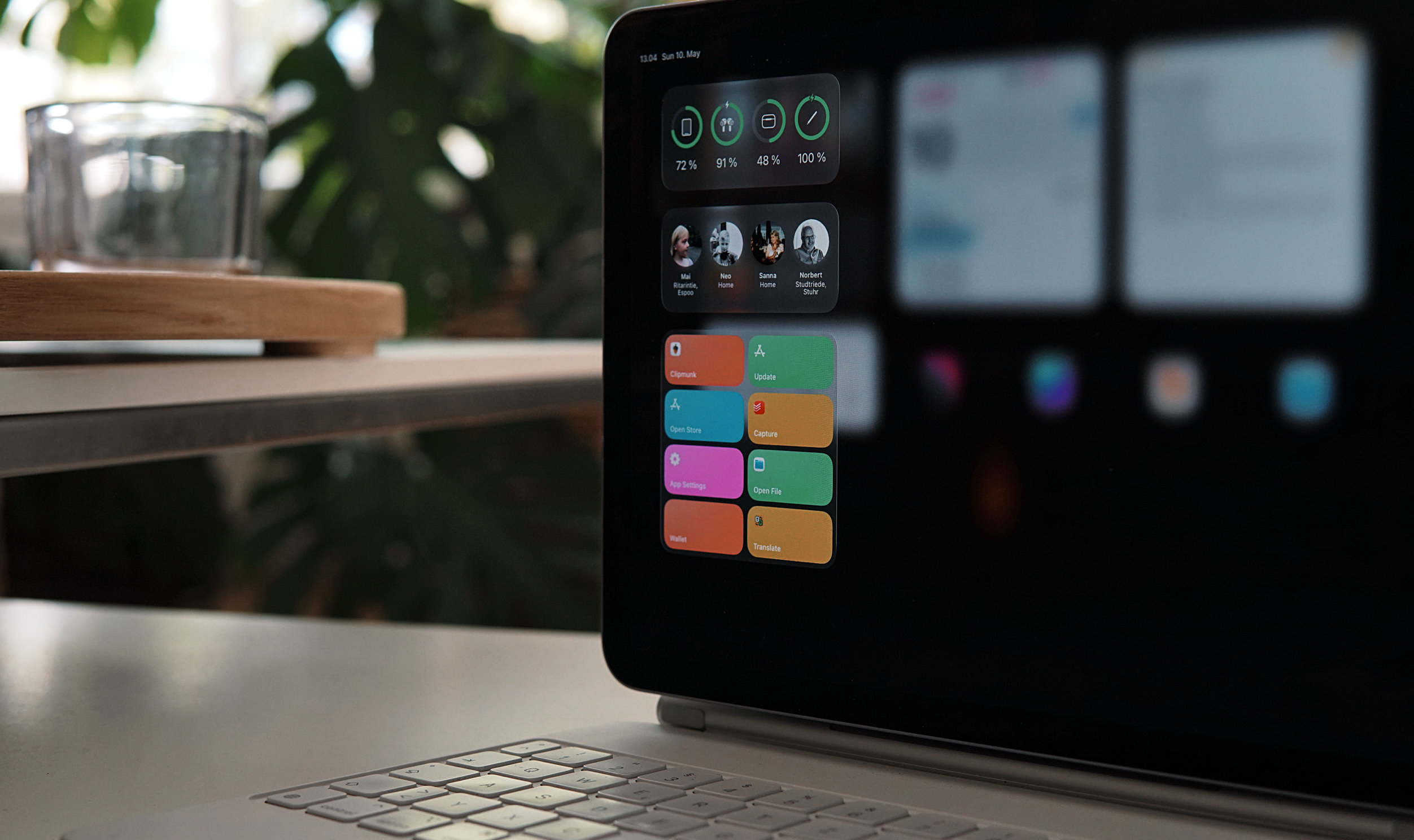Ein Tablet mit angeschlossener Tastatur auf einem Schreibtisch zeigt den iPadOS-Startbildschirm mit verschiedenen bunten Widgets und App-Symbolen, vor einem verschwommenen Hintergrund mit einer Pflanze und einem Glas.