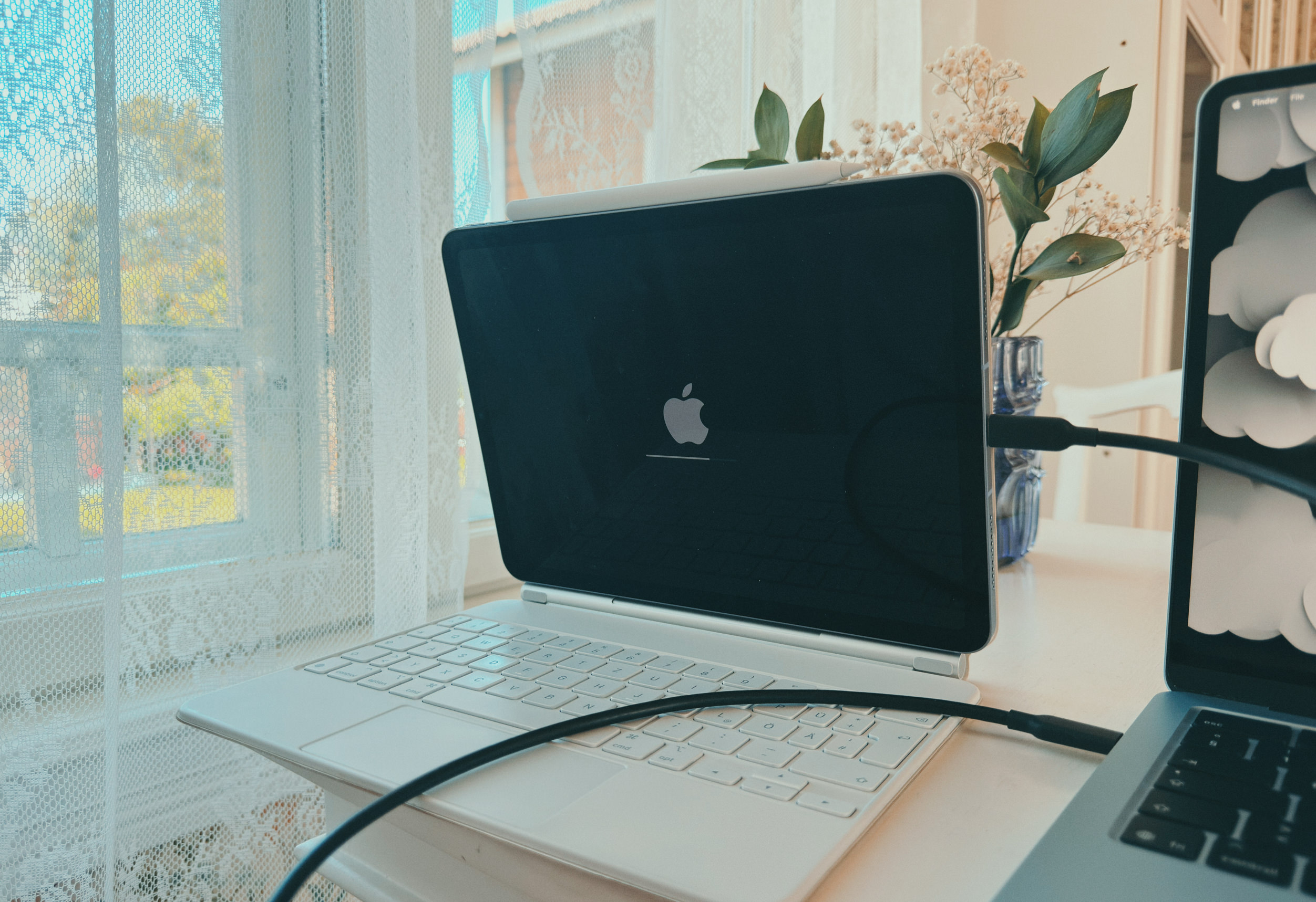 Ein Tablet in einer weißen Tastaturhülle steht auf einem Schreibtisch und zeigt das Apple-Logo auf einem schwarzen Bildschirm. Ein Ladekabel ist mit dem Gerät verbunden, das neben einem Laptop vor einem sonnigen Fenster mit Spitzengardinen steht.