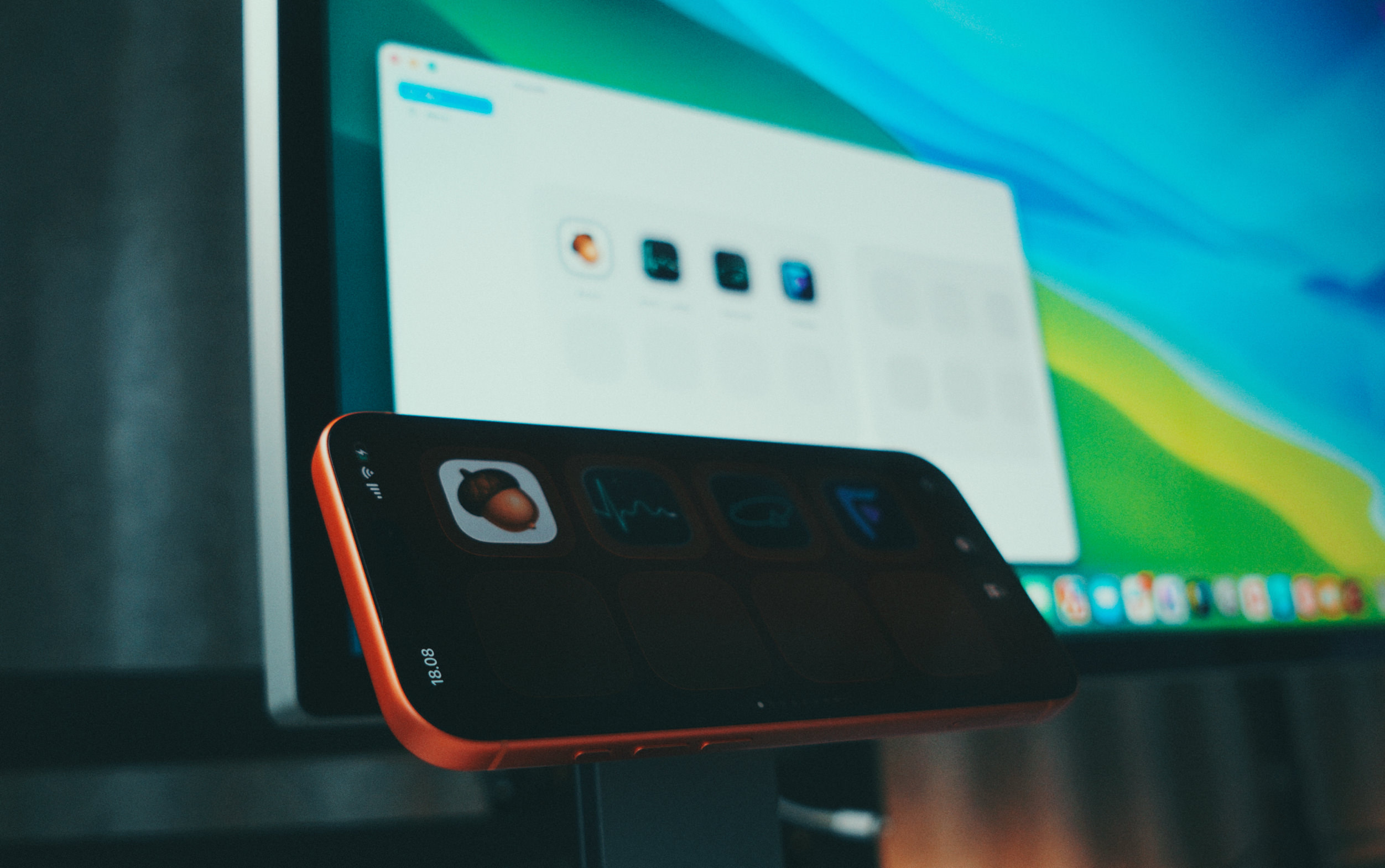A smartphone with an orange case is positioned in front of a computer monitor. The phone's screen displays several app icons, while the blurred background shows a macOS desktop interface.