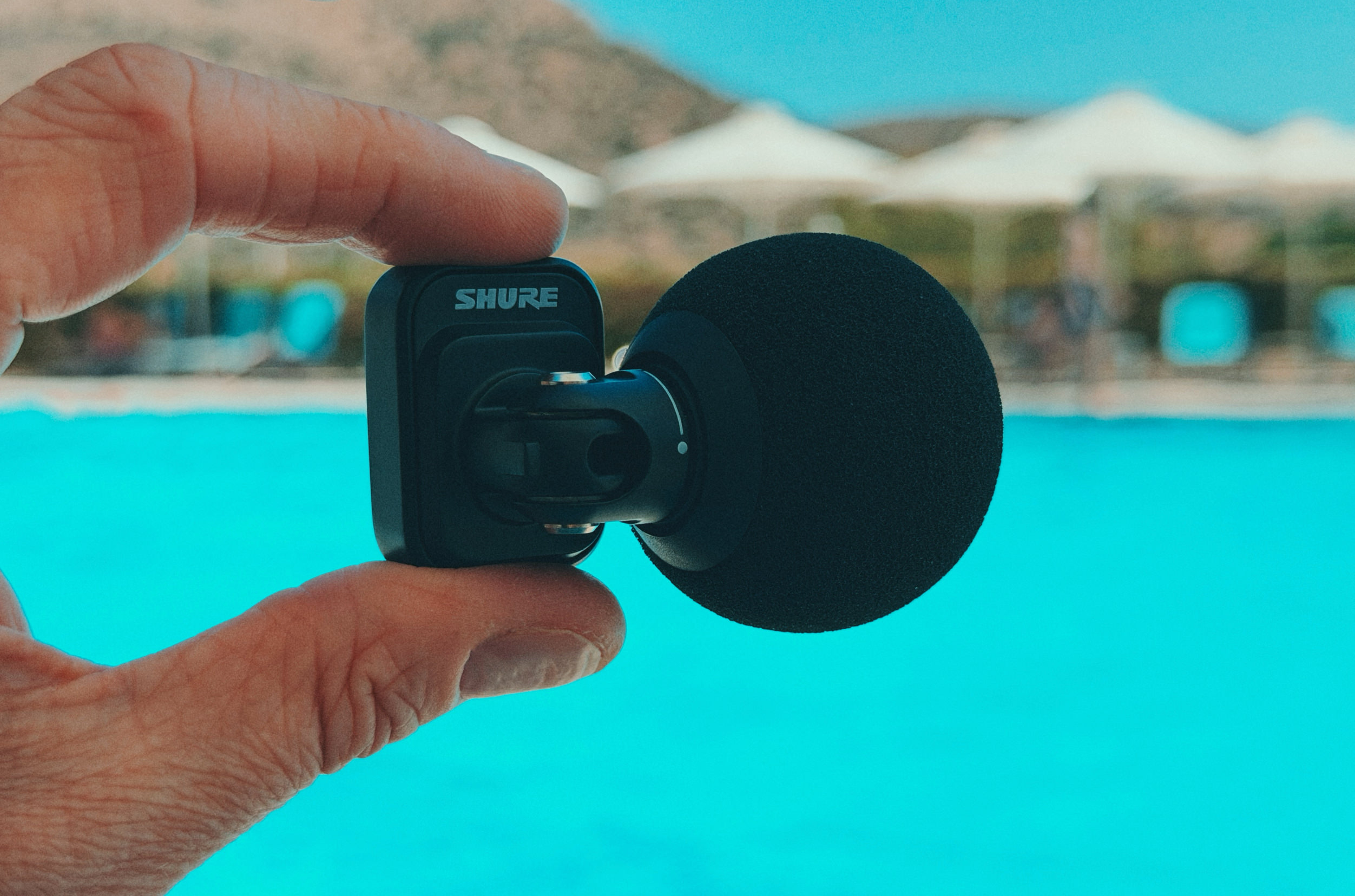 Eine Hand hält ein kleines Shure-Mikrofon mit einem runden schwarzen Schaumstoff-Windschutz vor einem helltürkisfarbenen Swimmingpool.