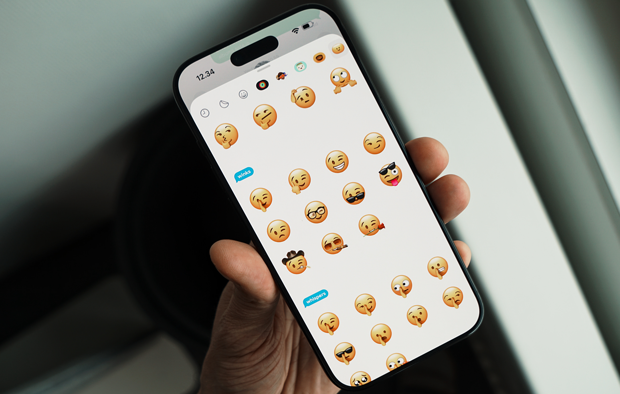 A hand holds a smartphone displaying an emoji selection screen, featuring various facial expressions and gestures. The phone's status bar shows the time as 12:34, indicating it is being used for communication or messaging.