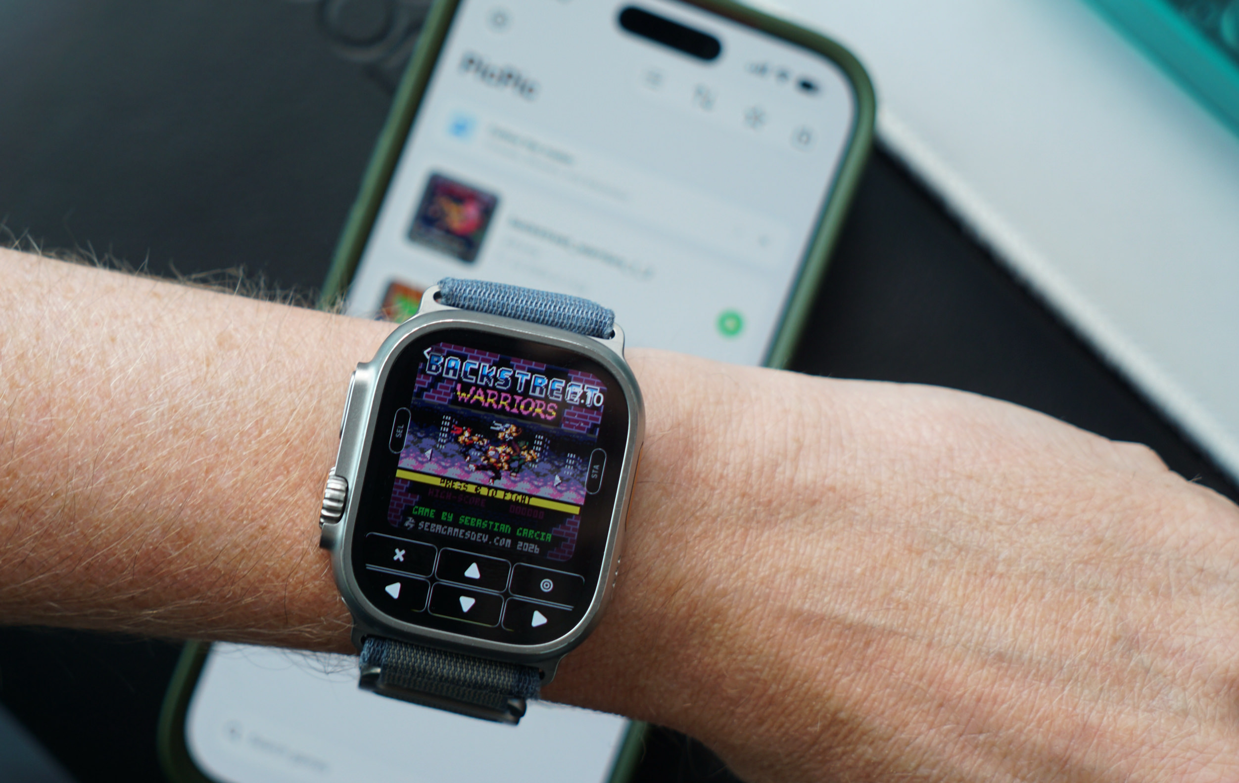 Eine Person trägt eine Smartwatch am Handgelenk, auf der ein Pixel-Art-Kampfspiel mit dem Titel „Backstreet Warriors“ und integrierter Spielsteuerung angezeigt wird, während im unscharfen Hintergrund ein Smartphone zu sehen ist.