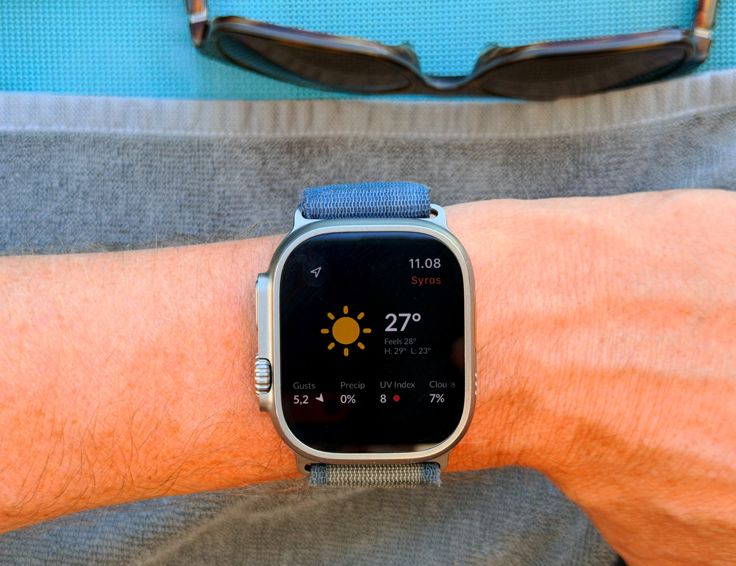 Eine Nahaufnahme einer Apple Watch Ultra am Handgelenk einer Person, die eine Wetter-App mit einer Temperatur von 27 Grad Celsius anzeigt, während das Handgelenk auf einem grauen Handtuch ruht und im Hintergrund eine Sonnenbrille zu sehen ist.