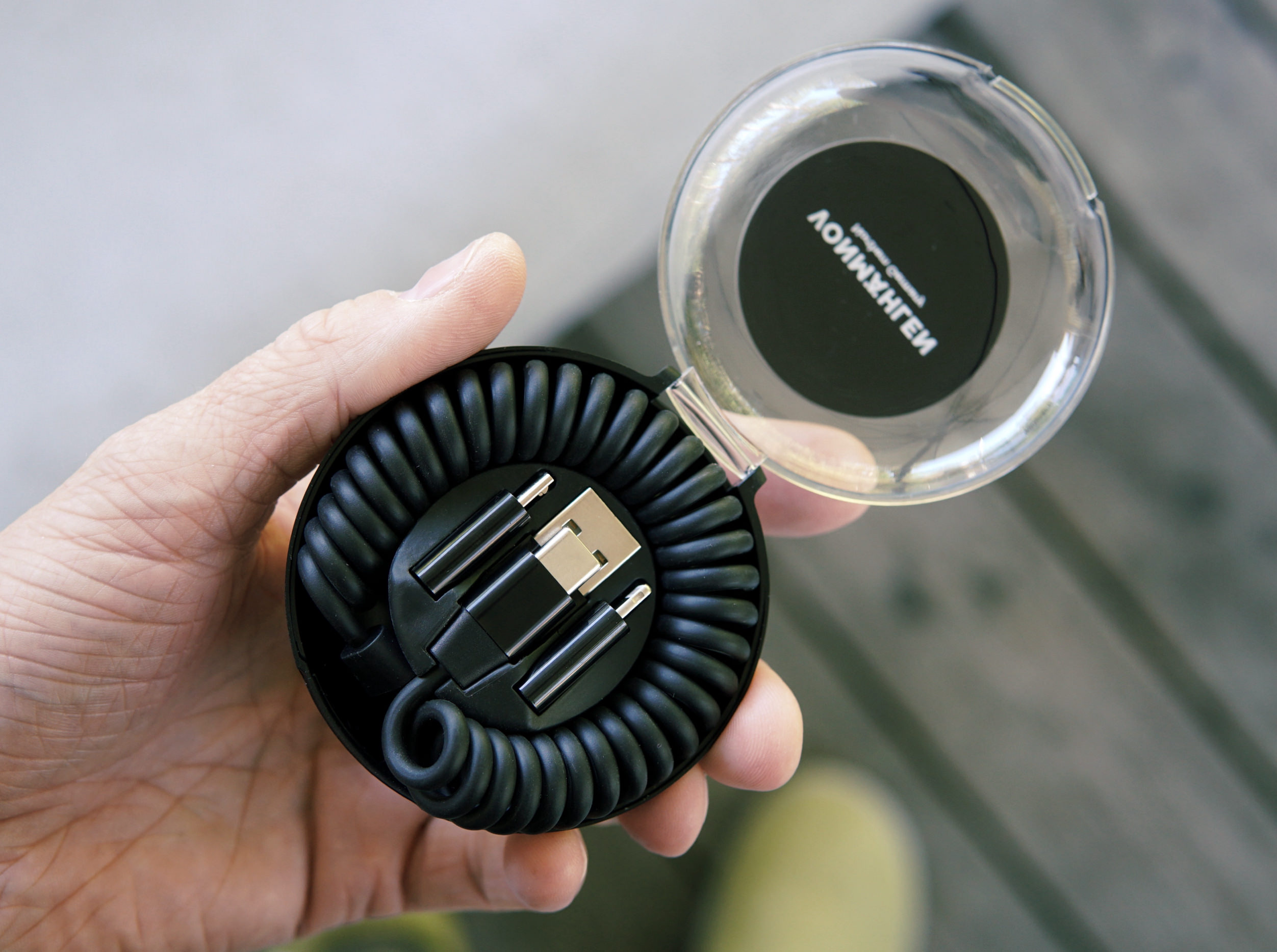 Eine Hand hält ein offenes, rundes schwarzes Etui, das ein schwarzes, spiralförmiges Multi-Adapter-Ladekabel enthält.