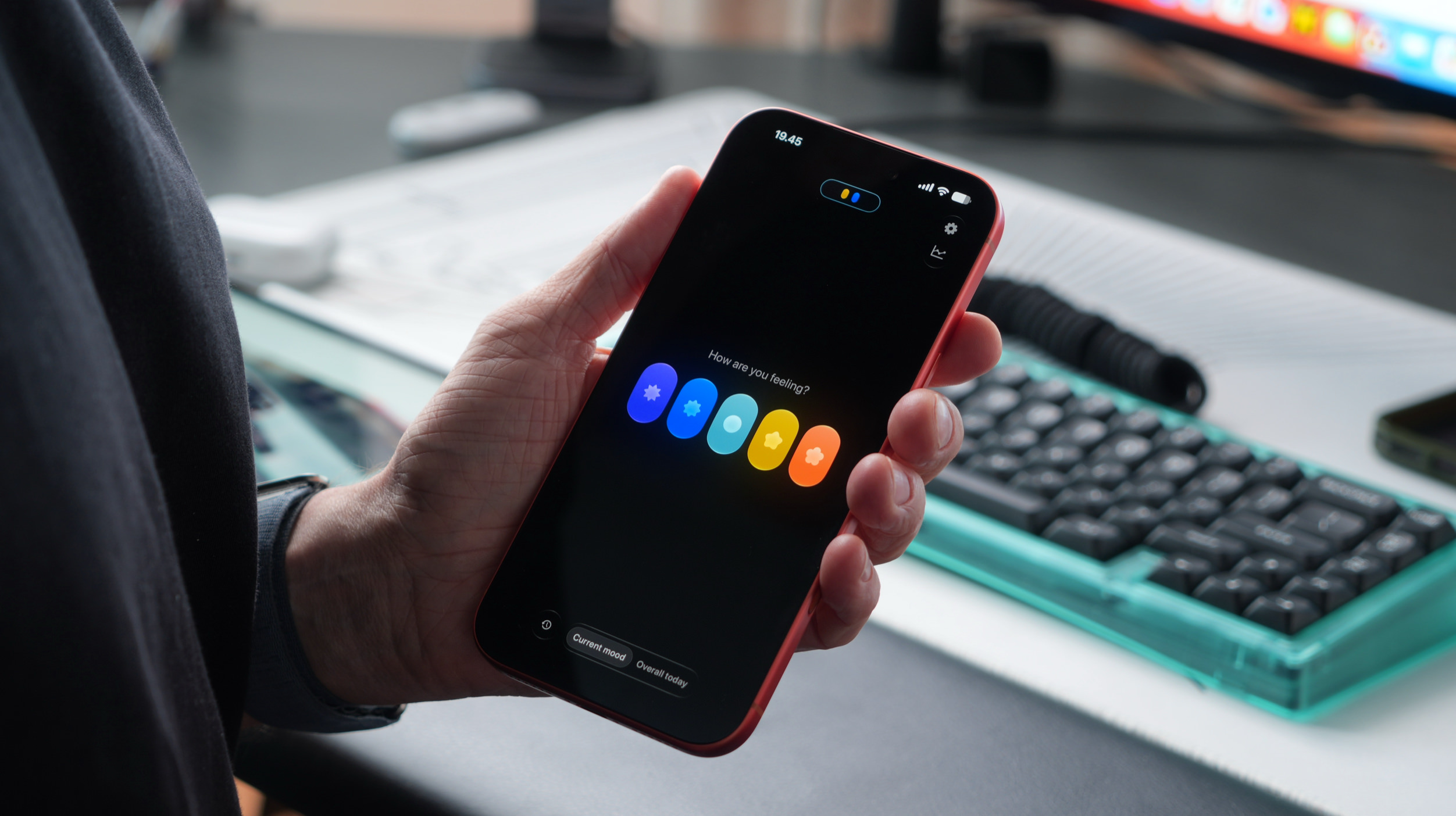 Eine Person hält ein Smartphone, auf dessen Display die Benutzeroberfläche einer Stimmungs-App mit fünf bunten, blumenförmigen Symbolen unter der Frage „Wie fühlst du dich?“ in einer Büroumgebung angezeigt wird.