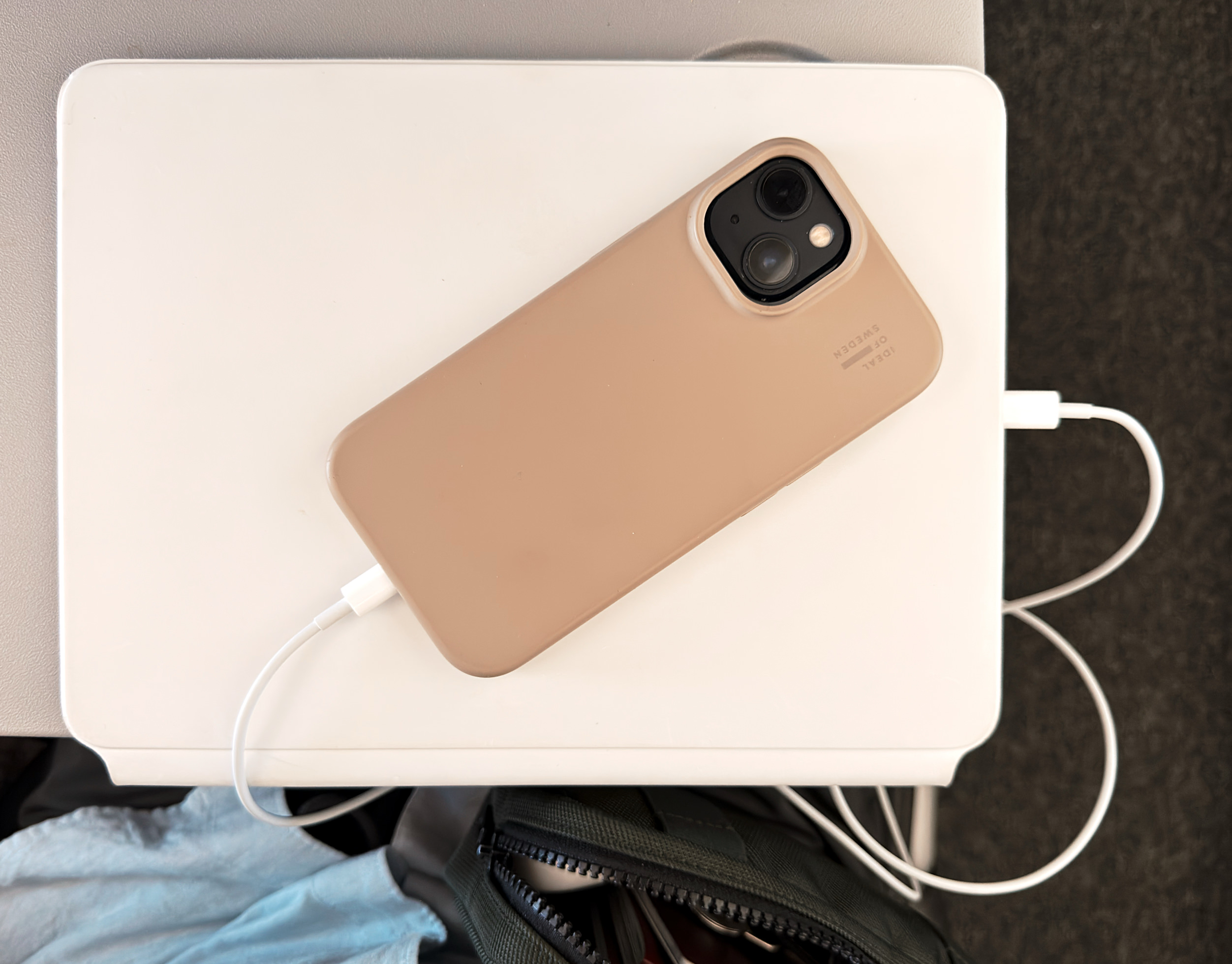 Ein beigefarbenes Smartphone in einer Schutzhülle liegt auf einem flachen weißen Tablet oder Laptop, an das ein weißes Ladekabel angeschlossen ist. Darunter ist eine Tasche teilweise zu sehen.