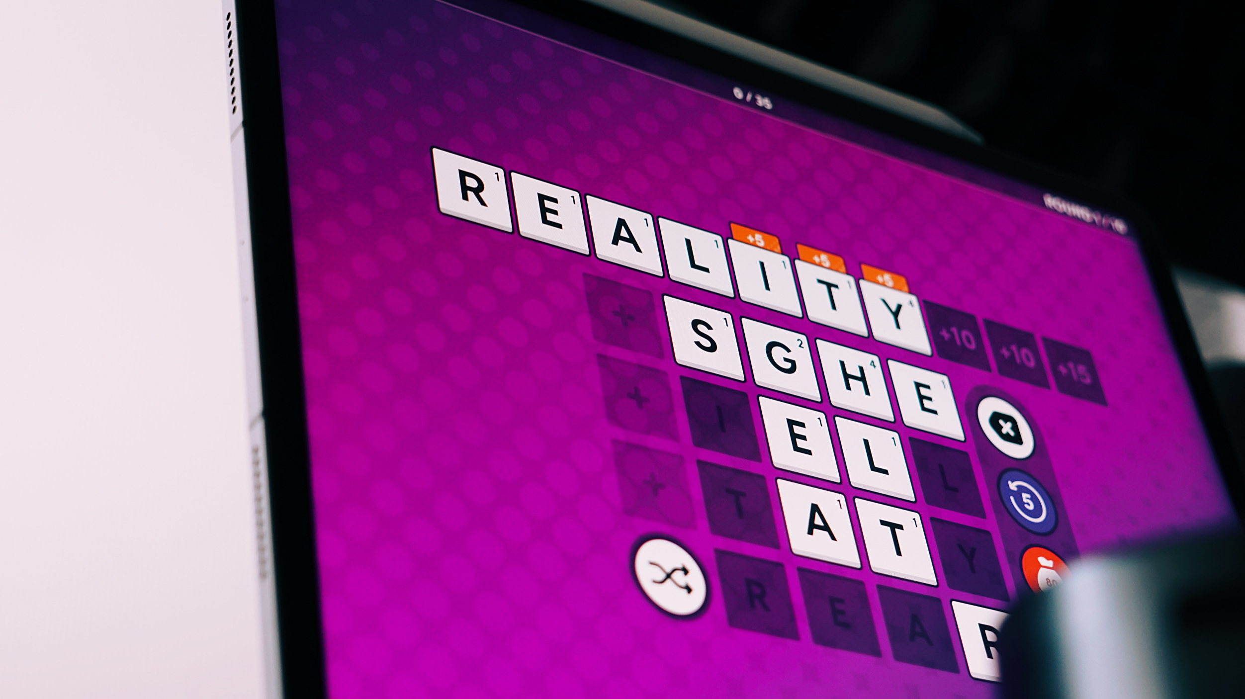 Ein Tablet-Bildschirm zeigt ein mobiles Worträtselspiel mit dem Wort „REALITY“, das in einem Kreuzworträtsel-Gitter auf einem lilafarbenen Hintergrund vervollständigt wurde.