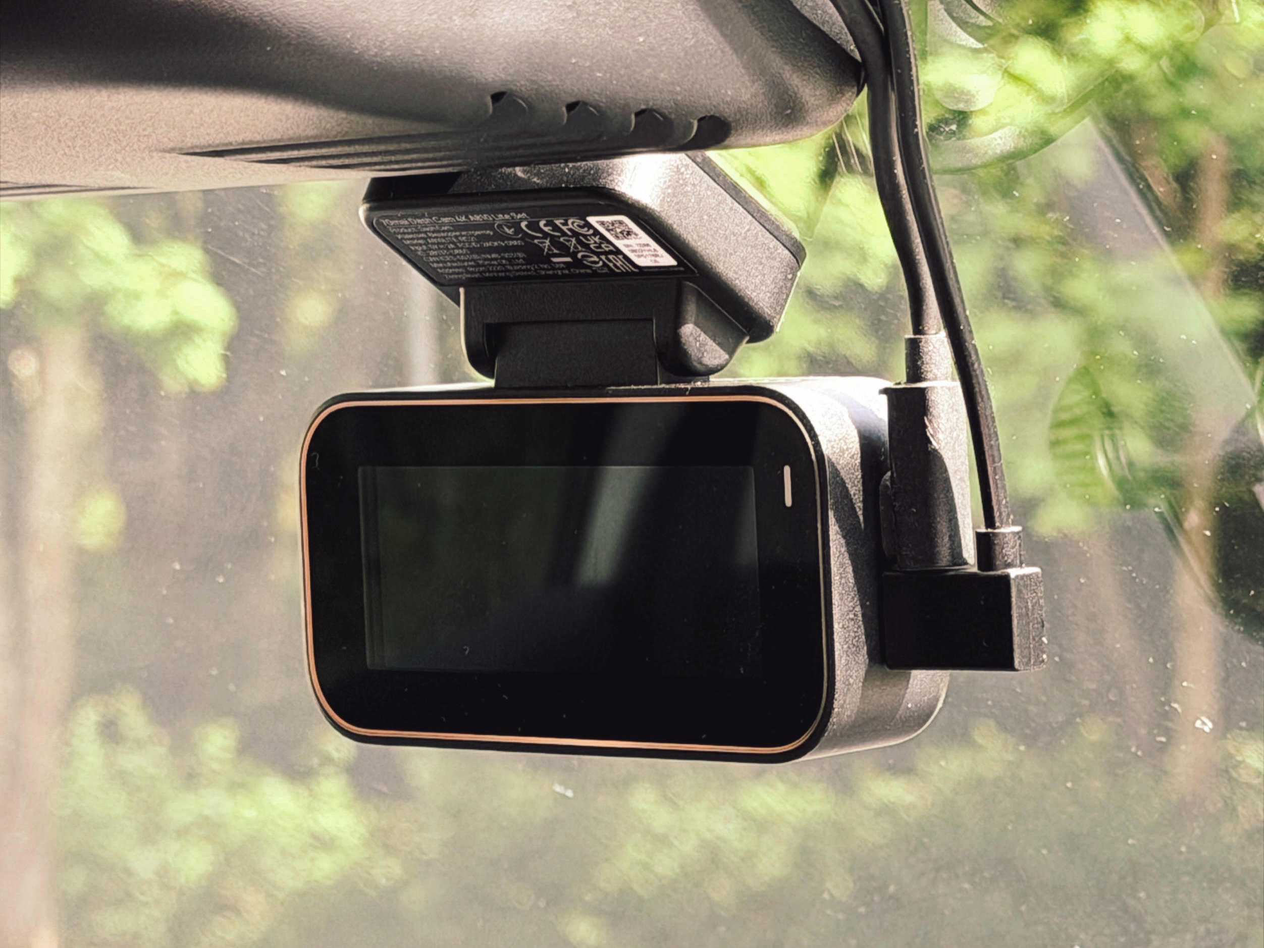 Eine schwarze Dashcam mit kupferfarbenem Rand, die an der Innenseite einer Windschutzscheibe montiert ist, mit angeschlossenem Stromkabel und verschwommenen grünen Bäumen im Hintergrund.