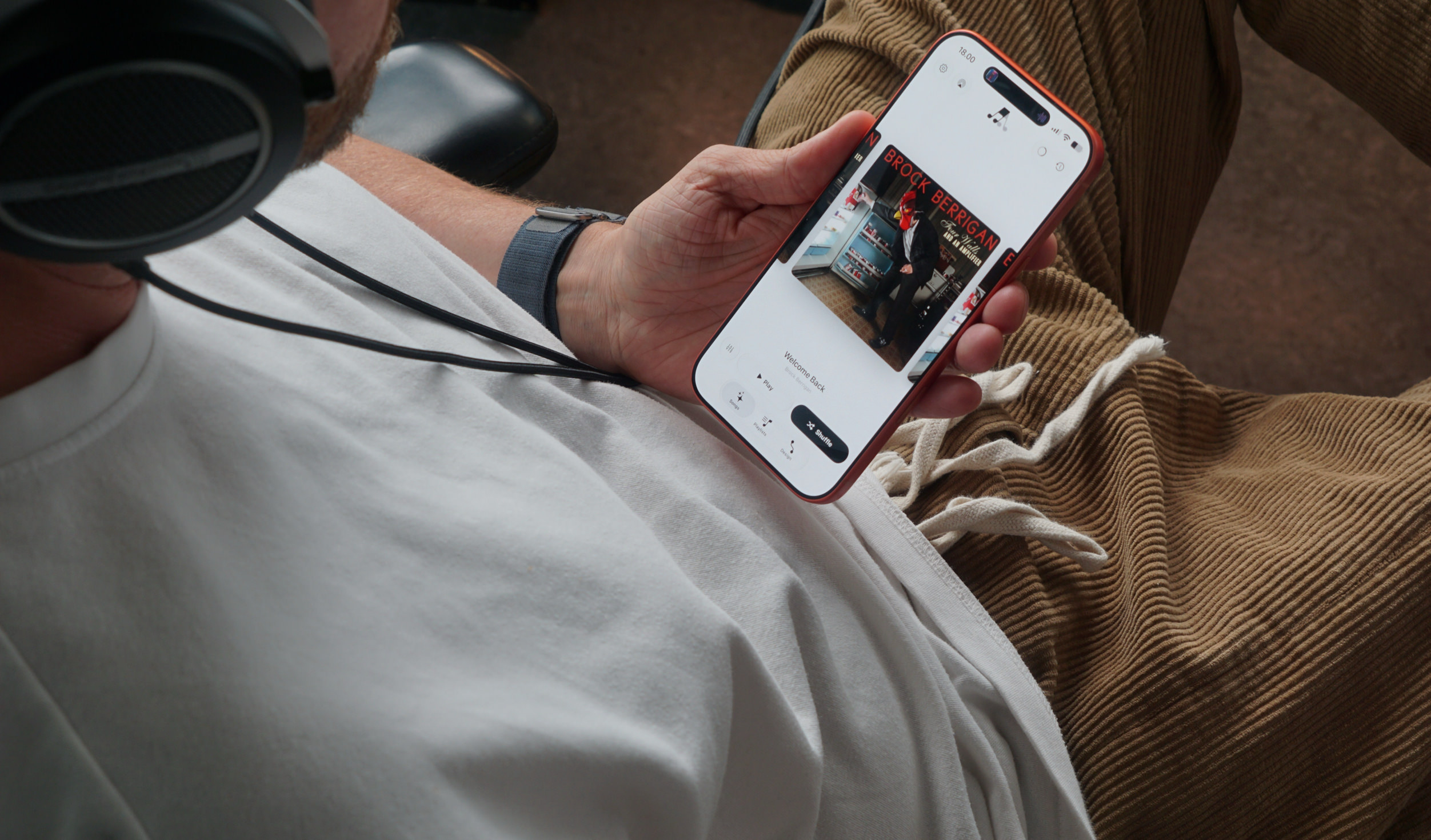 Eine Person in einem weißen T-Shirt, einer beigen Cordhose und Kopfhörern hält ein Smartphone mit einer Musik-App-Oberfläche in der Hand.
