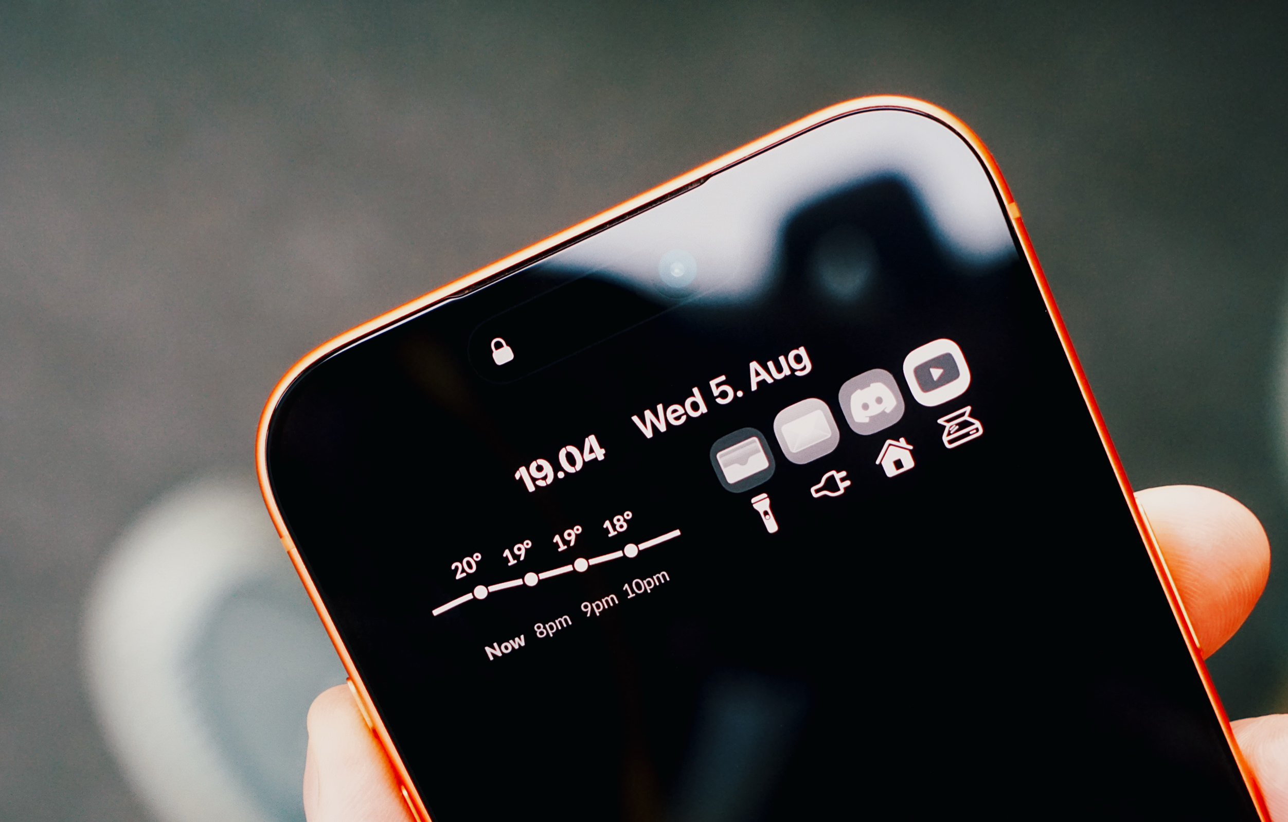 Nahaufnahme eines orangefarbenen Smartphones in einer Hand. Der Sperrbildschirm im dunklen Design zeigt die Uhrzeit, das Datum, ein Wettervorhersage-Diagramm sowie eine Anordnung verschiedener App-Symbole.