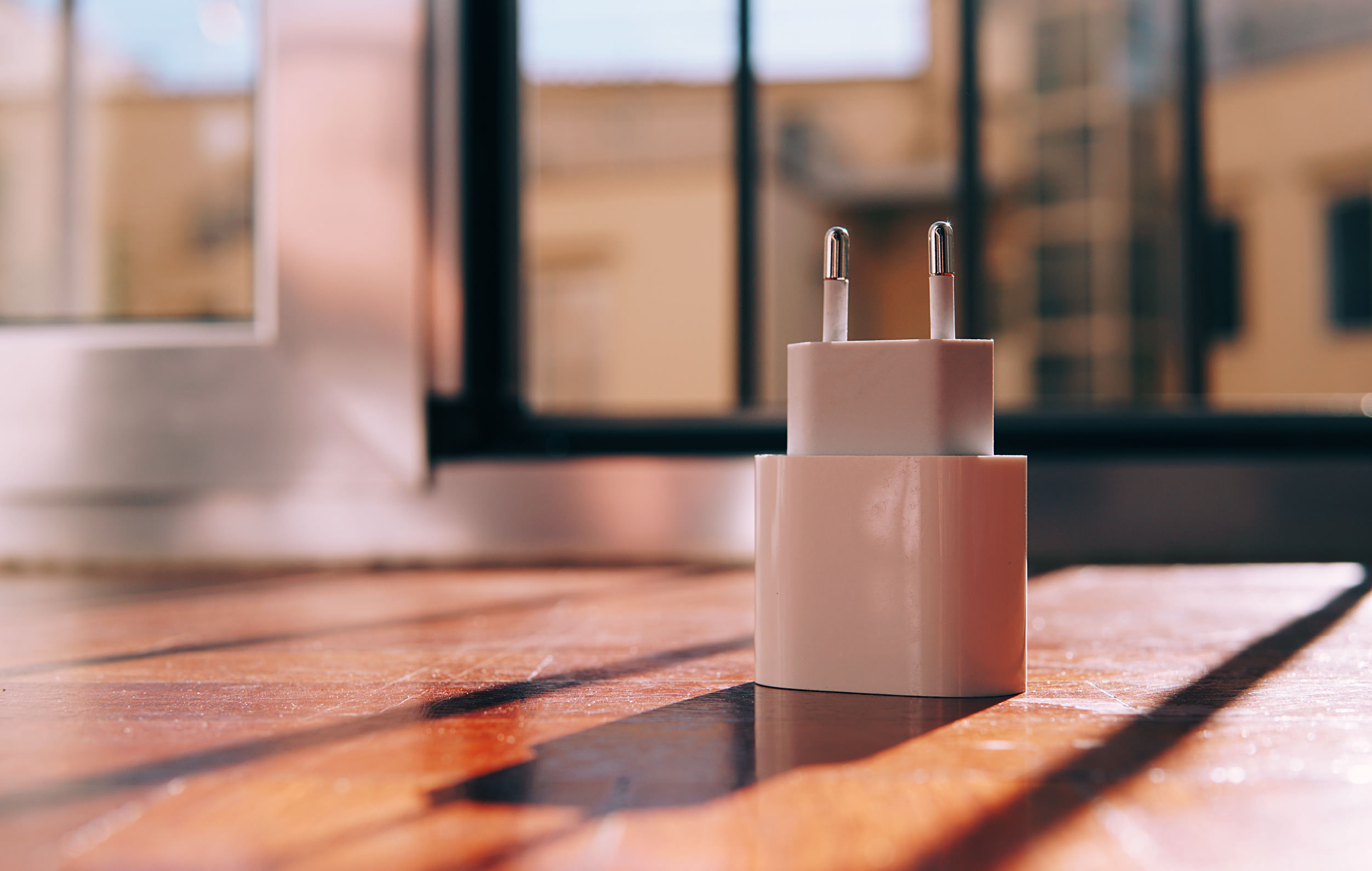 A close-up photograph of a white power adapter resting on a wooden table in front of a window.