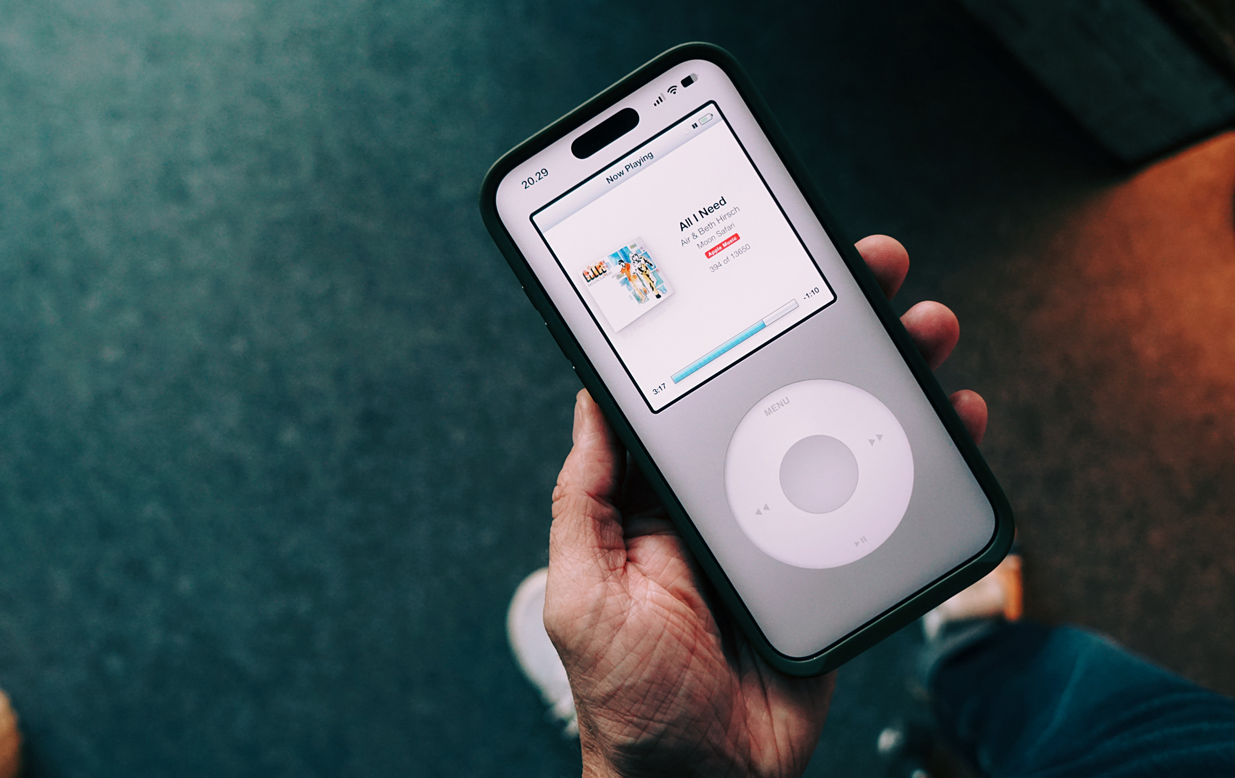 Eine Nahaufnahme einer Hand, die ein Smartphone hält, auf dessen Display eine Musik-App im Stil eines klassischen iPods mit Click-Wheel angezeigt wird.
