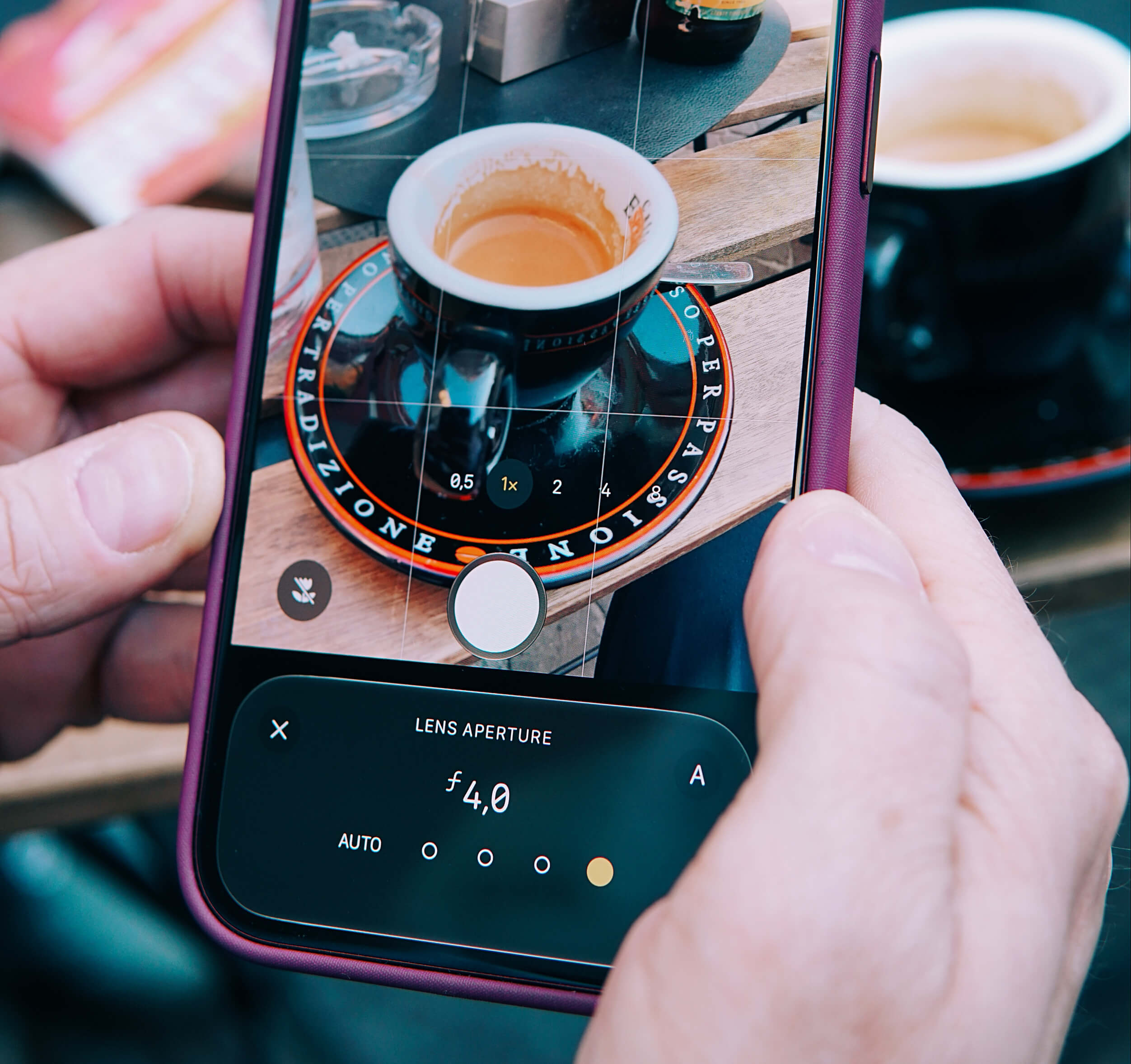 A close-up of a person holding a smartphone taking a photo of a small coffee cup on a saucer placed on a wooden table. The phone screen shows camera settings including a lens aperture of f4.0, and the focus is on the coffee cup in the center of the frame.