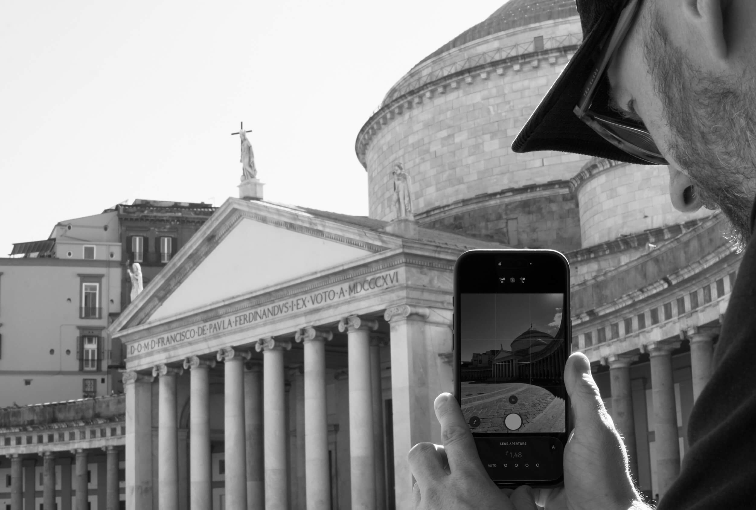 A man wearing glasses and a hat is holding a smartphone and taking a photo of a classical building with columns and statues on the roof. The building has a triangular pediment with an inscription and a large dome behind it.