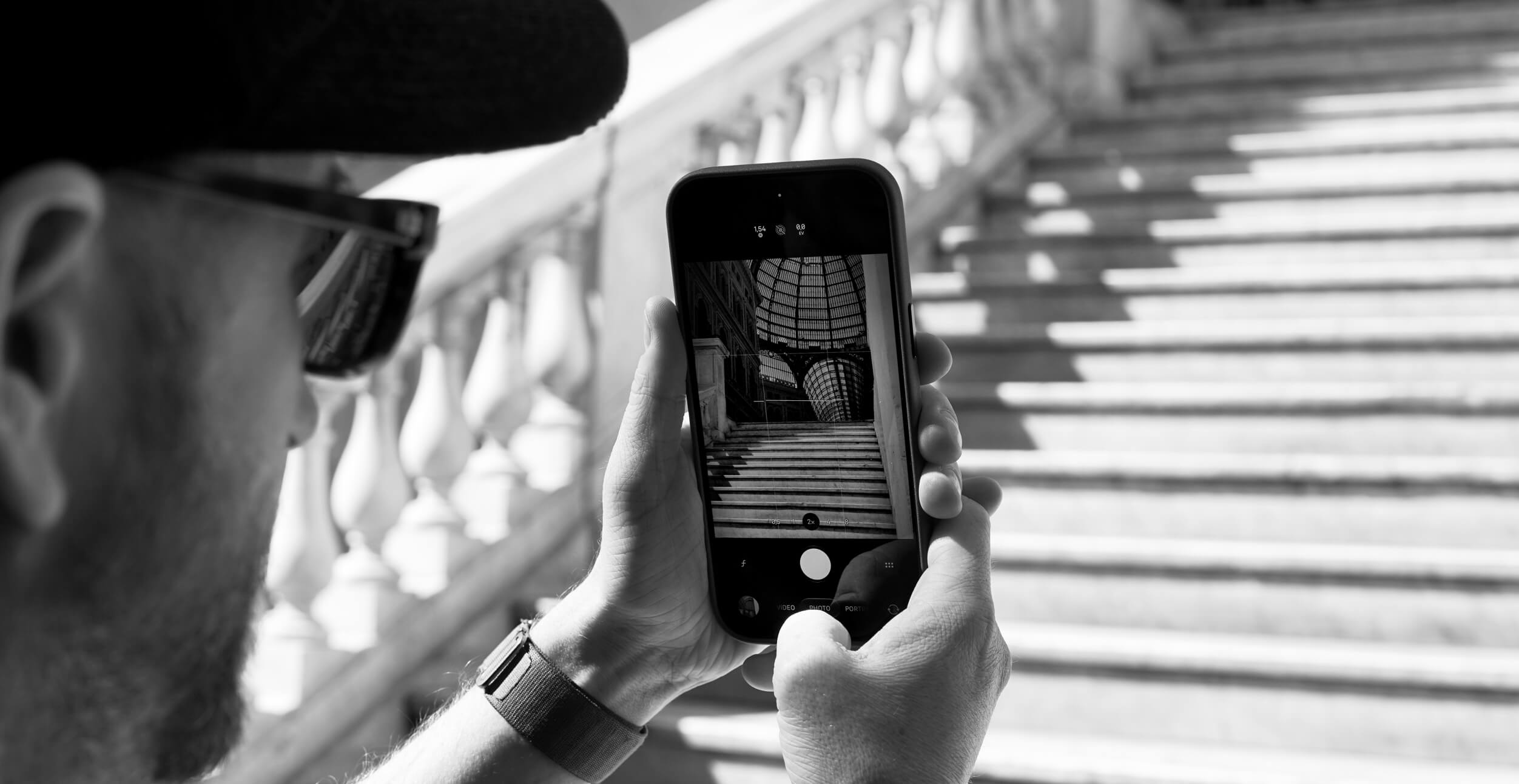 A person wearing a cap and sunglasses is holding a smartphone with both hands and taking a black and white photo of a staircase with balustrades and a glass-domed ceiling. The image on the phone screen shows the staircase and architectural details clearly.