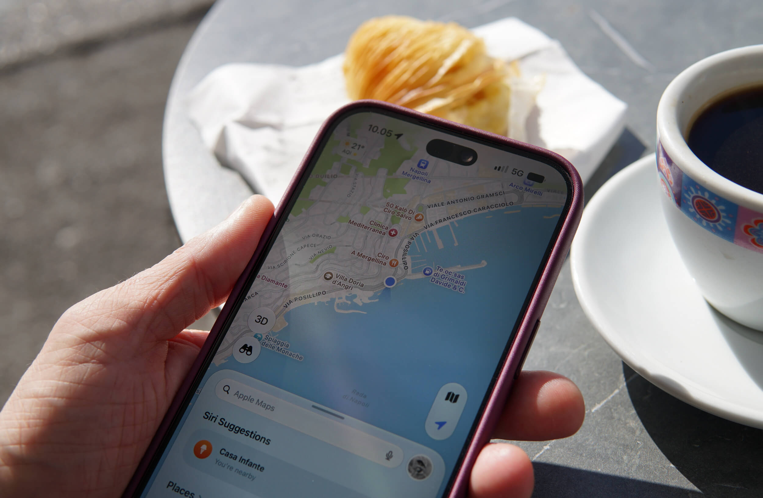 A hand holding a smartphone displaying a map application of a coastal area, with a plate of pastry and a cup of black coffee on a gray table surface nearby.